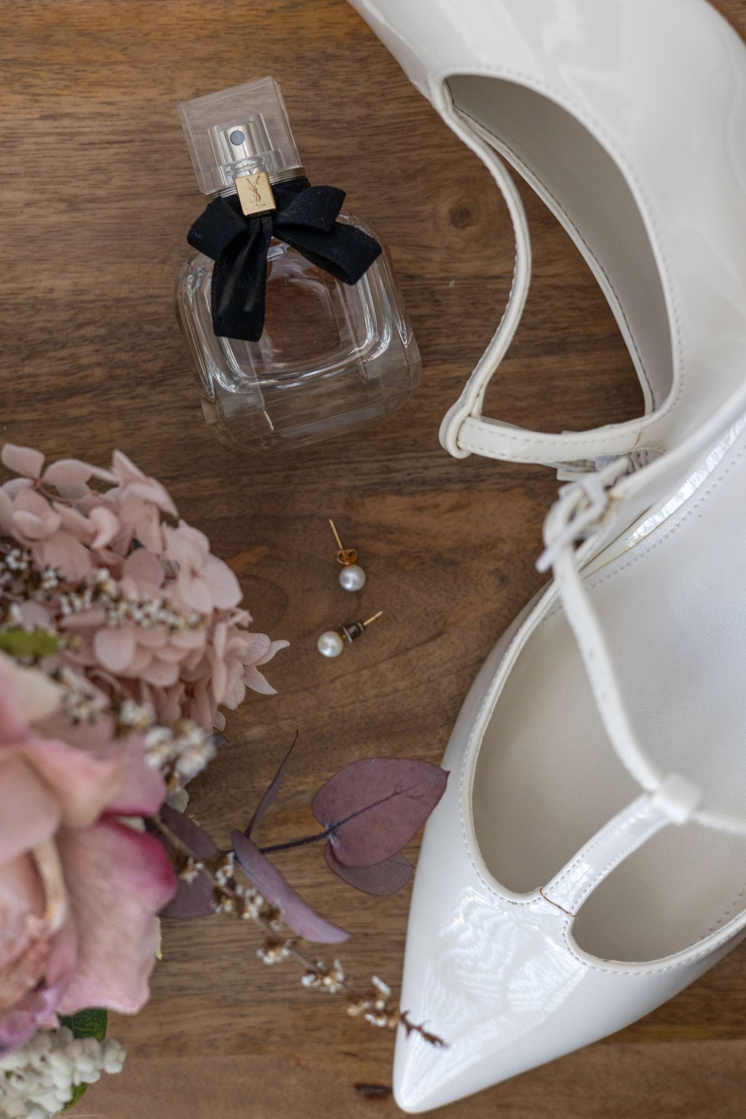 Bridal flat lay with white patent heels, pink floral bouquet, pearl earrings, and perfume bottle on wood.