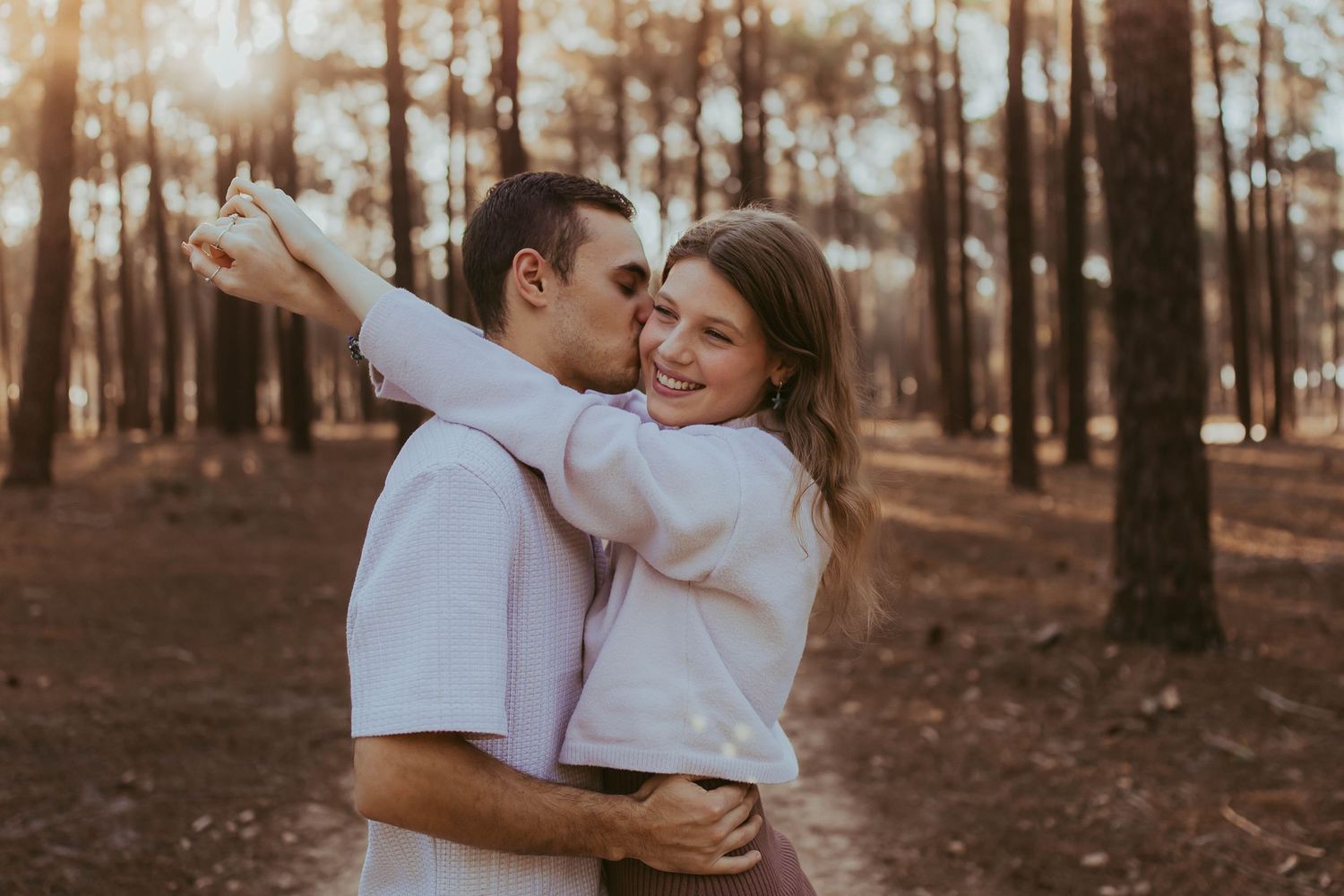 Couple embracing and sharing a kiss in a sunlit pine forest during golden hour.