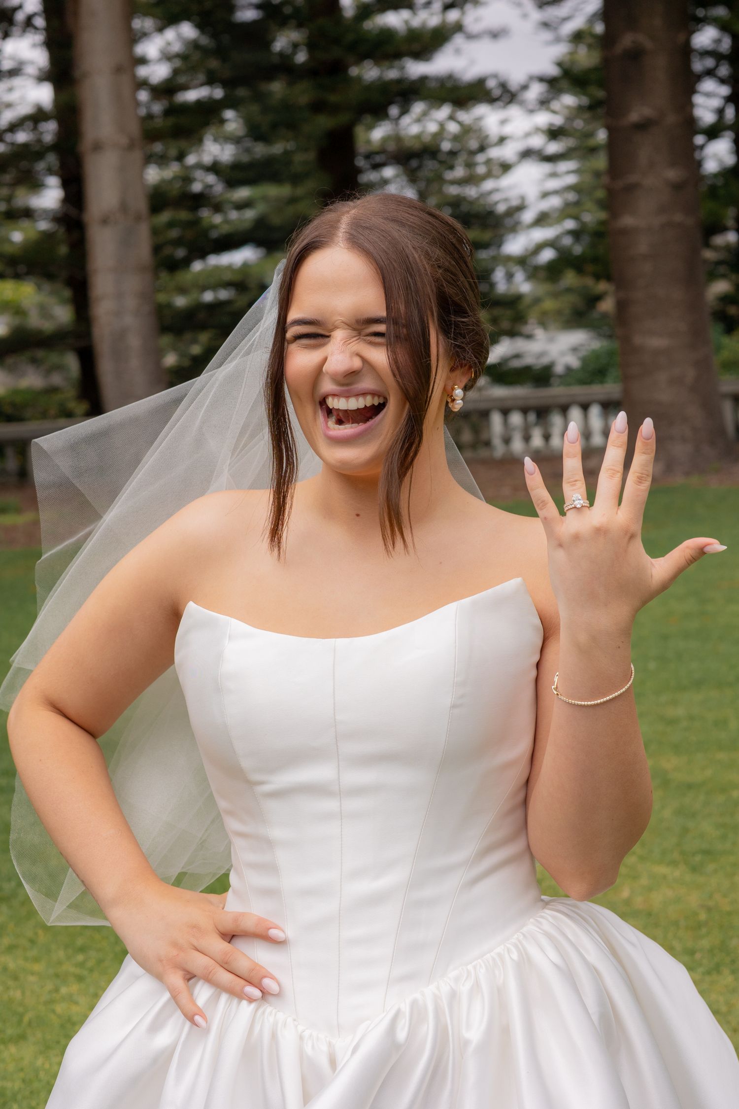 A joyful bride in a sleeveless white wedding dress laughs and poses outdoors against a backdrop of trees.