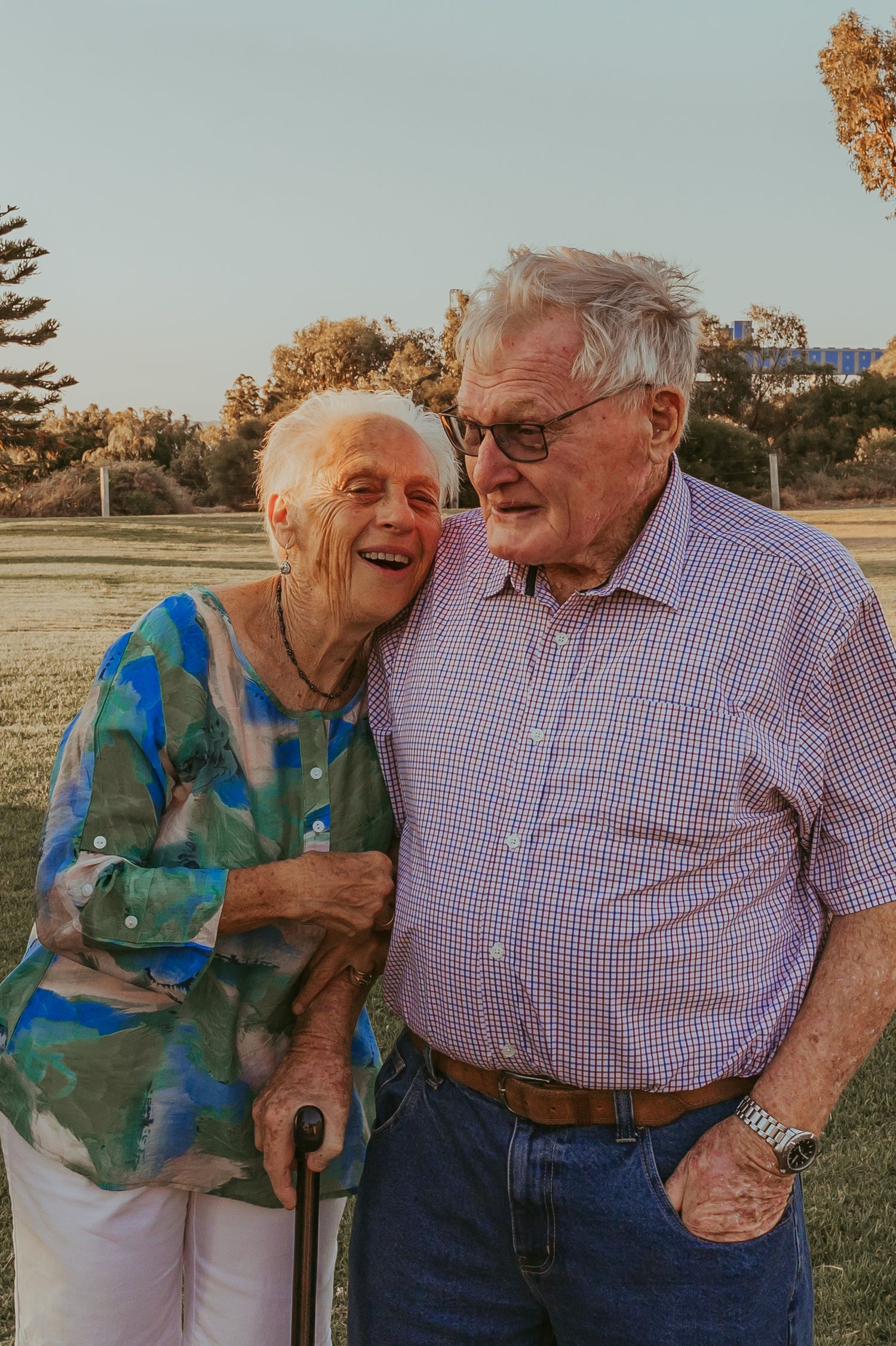An elderly couple laughing together outdoors during golden hour, wearing casual clothes and sharing a joyful moment.