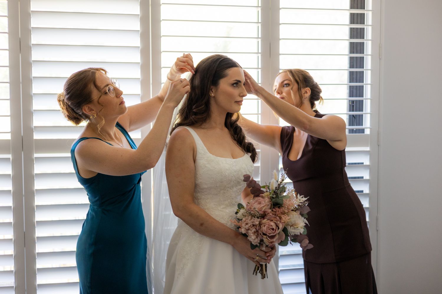 Bride in white gown holds bouquet while two women in blue and burgundy dresses adjust her hair before wedding ceremony.