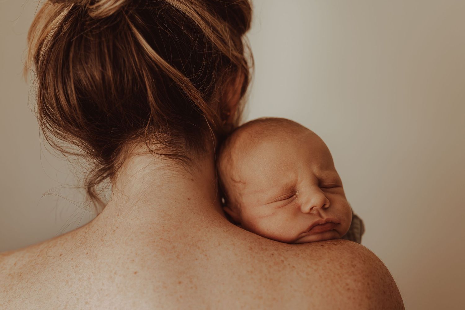 A newborn baby sleeping peacefully on a mother's bare freckled shoulder, captured in soft natural light.