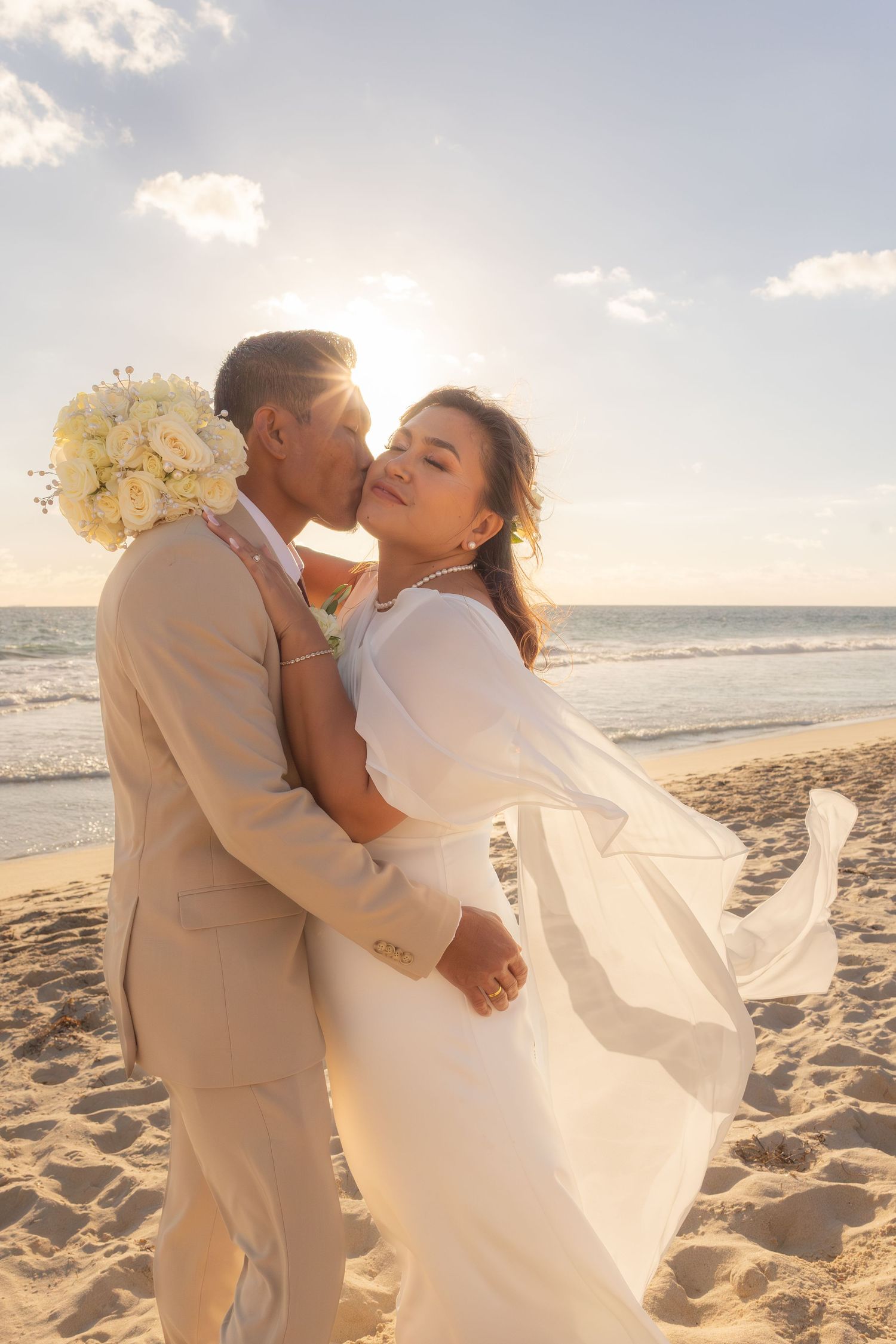 Newlywed couple sharing a romantic kiss on a sandy beach at sunset, bride in white gown holding bouquet.