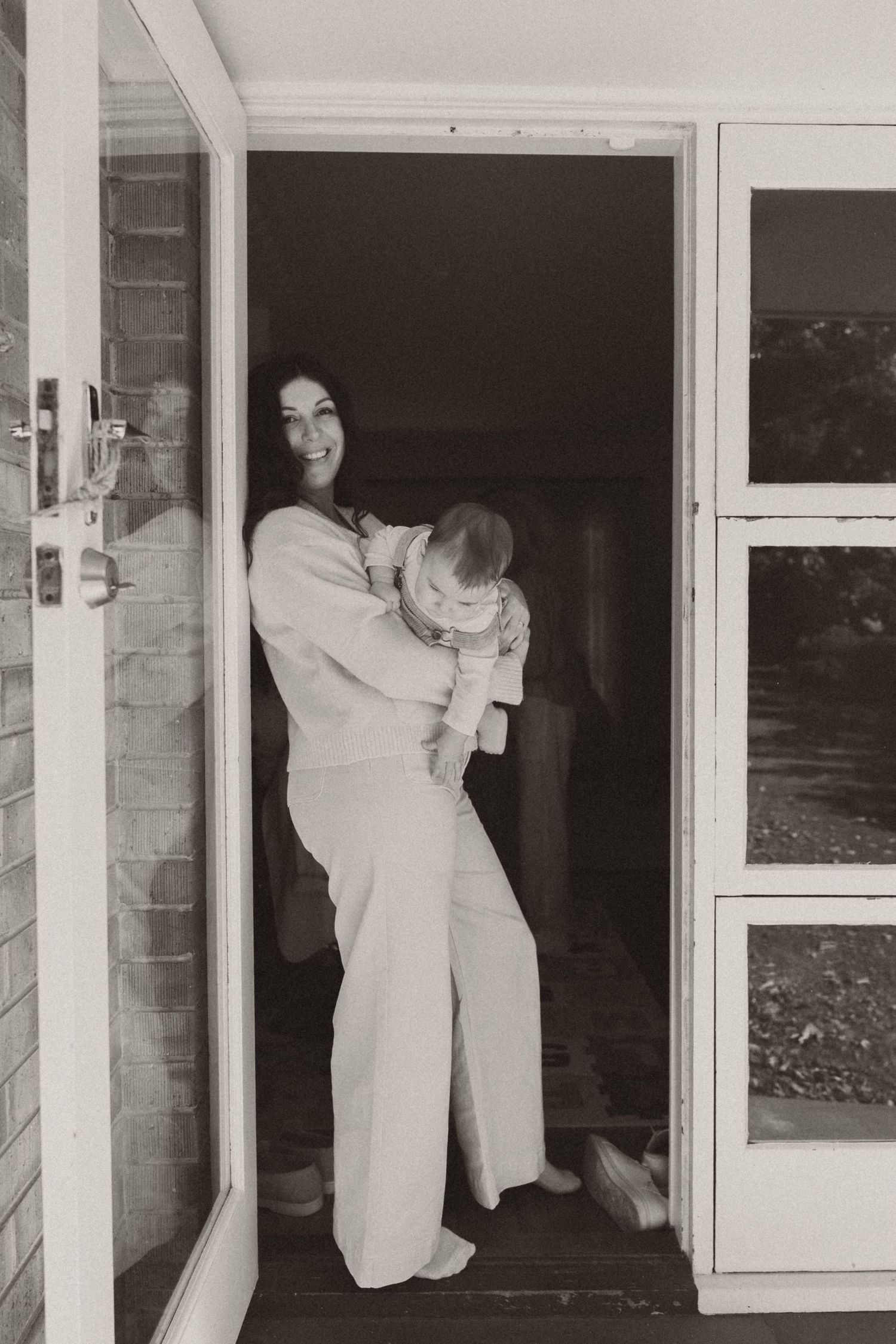 Black and white photo of a smiling woman holding a baby while standing in an open doorway of a home.