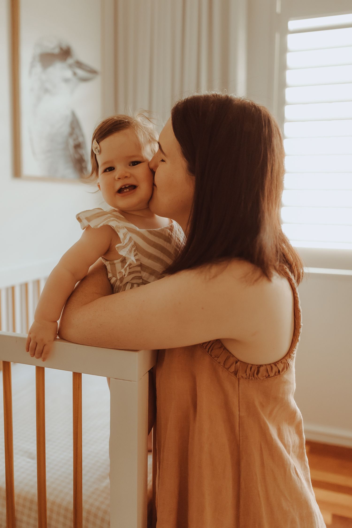 A mother in a tan dress holds and plays with her baby in a sunlit room.