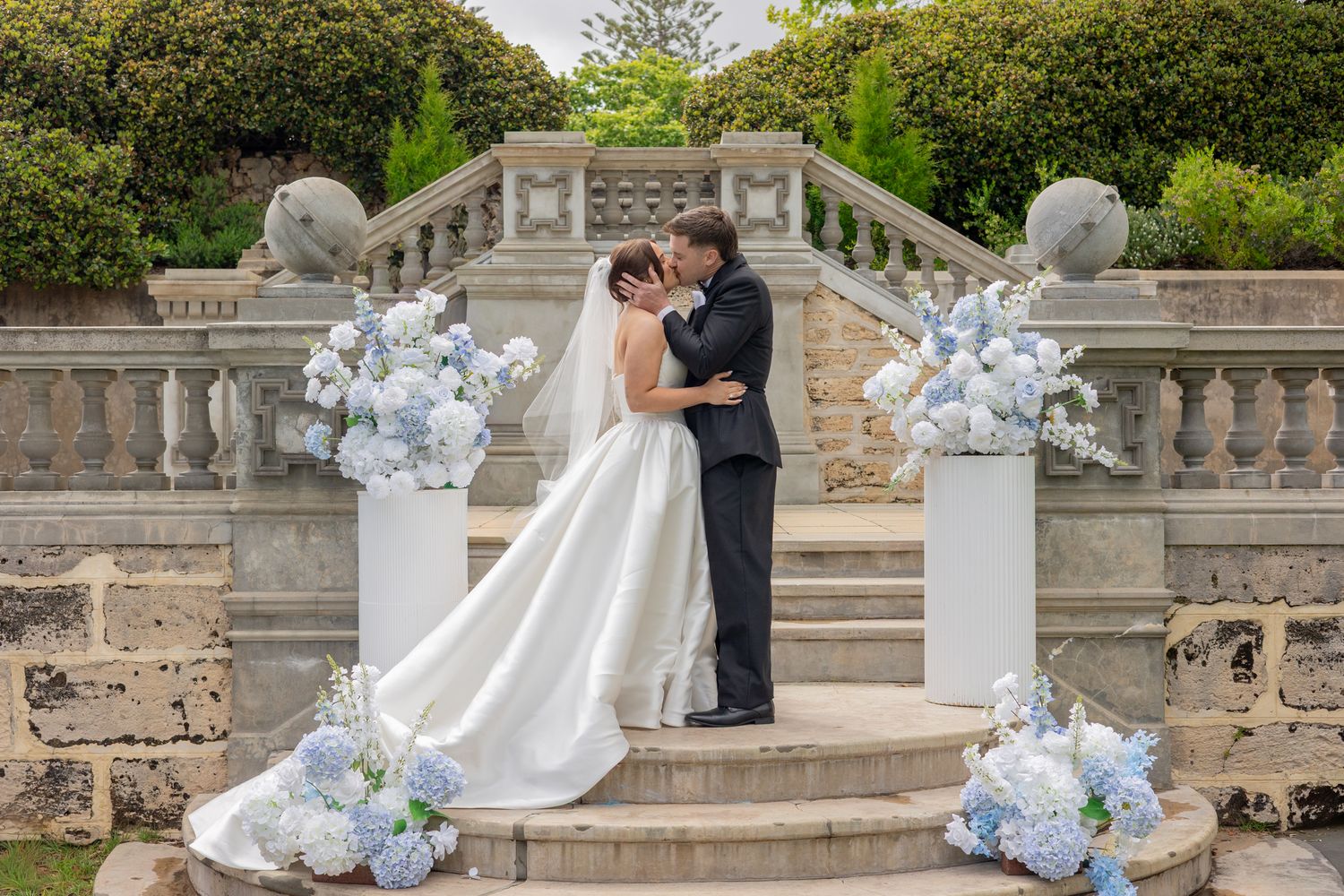 A couple shares an intimate moment on grand stone steps adorned with elegant blue and white floral arrangements.