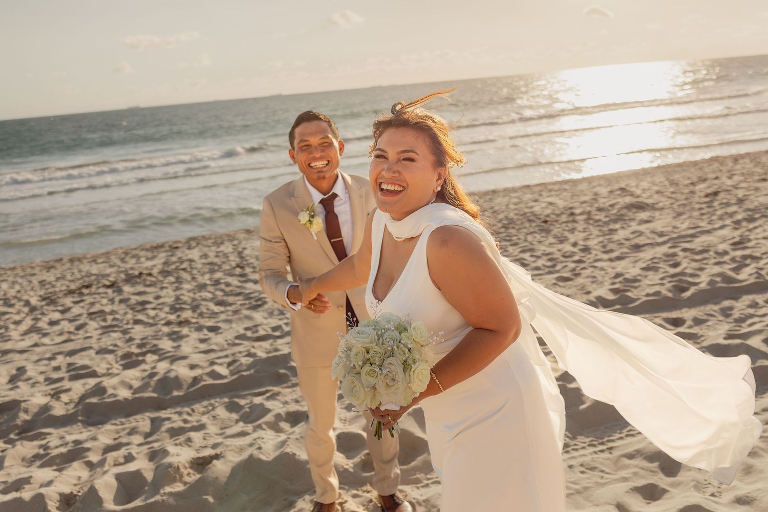 Joyful bride in white dress holding bouquet with groom in tan suit celebrating on a sunny beach wedding day.