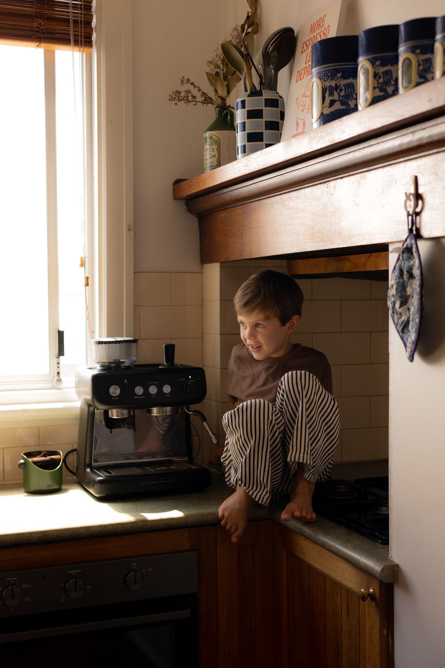 Child in striped shirt looking out kitchen window in warm sunlit interior space.