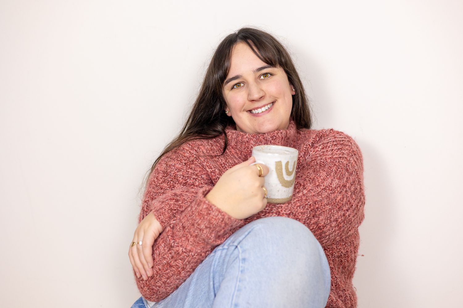 Person wearing a cozy pink sweater and light blue jeans holds a white coffee mug while smiling at the camera.