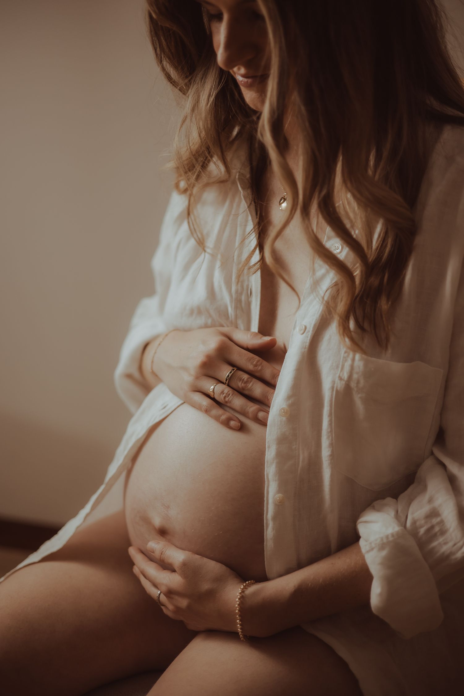 Intimate maternity photo of woman in white shirt cradling pregnant belly in soft warm lighting.