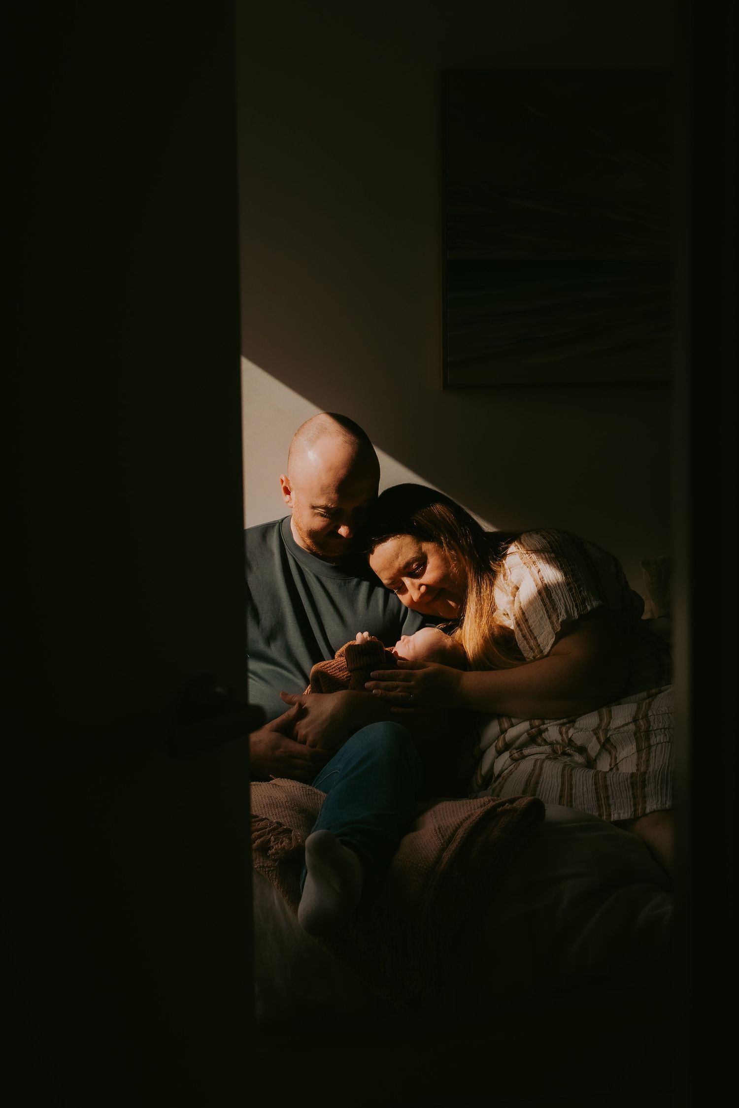A couple sits in soft light, tenderly cradling their newborn baby together in a warmly lit room.