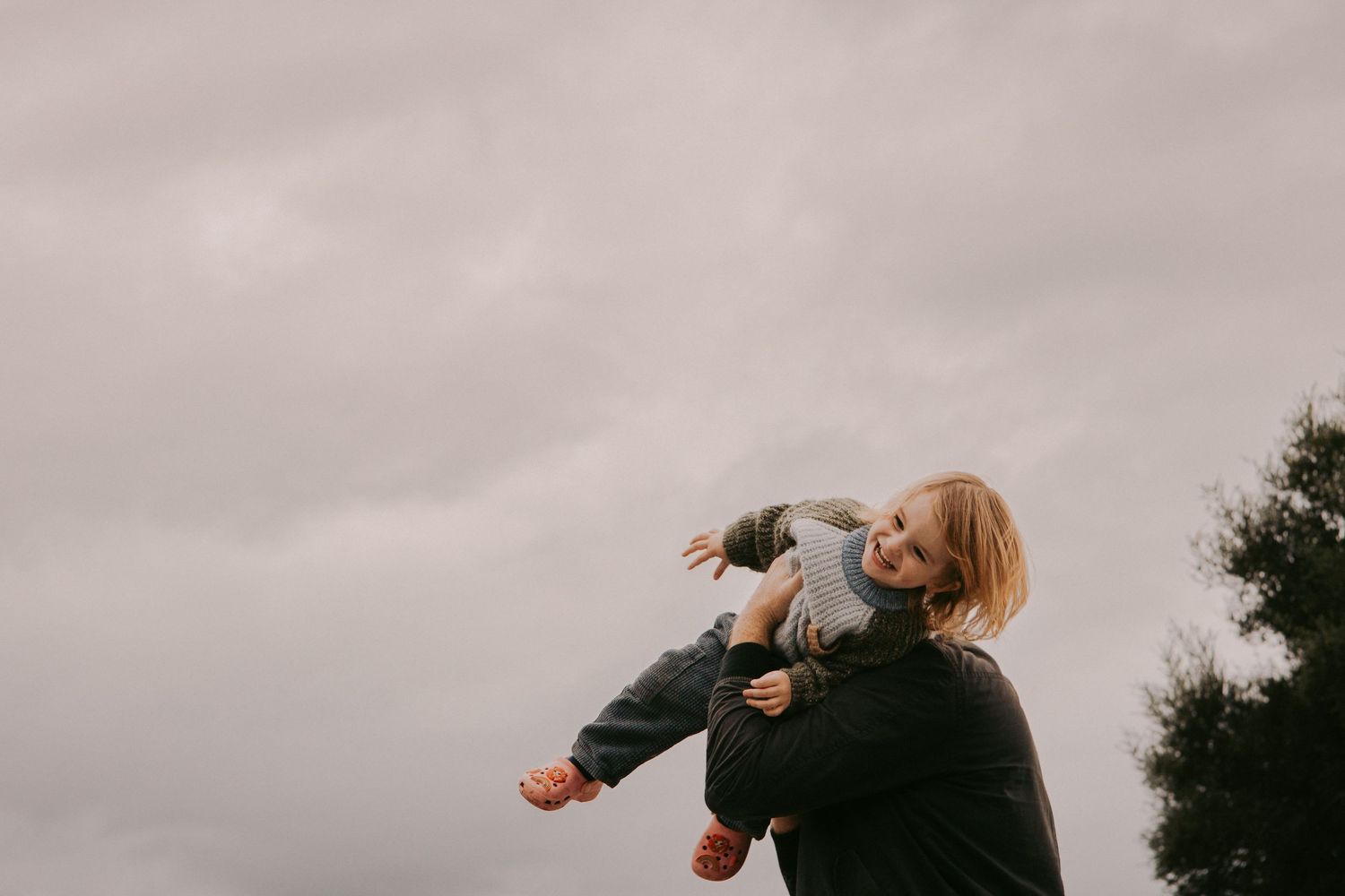 Parent joyfully lifting laughing child outdoors under overcast sky, capturing a playful and loving family moment.