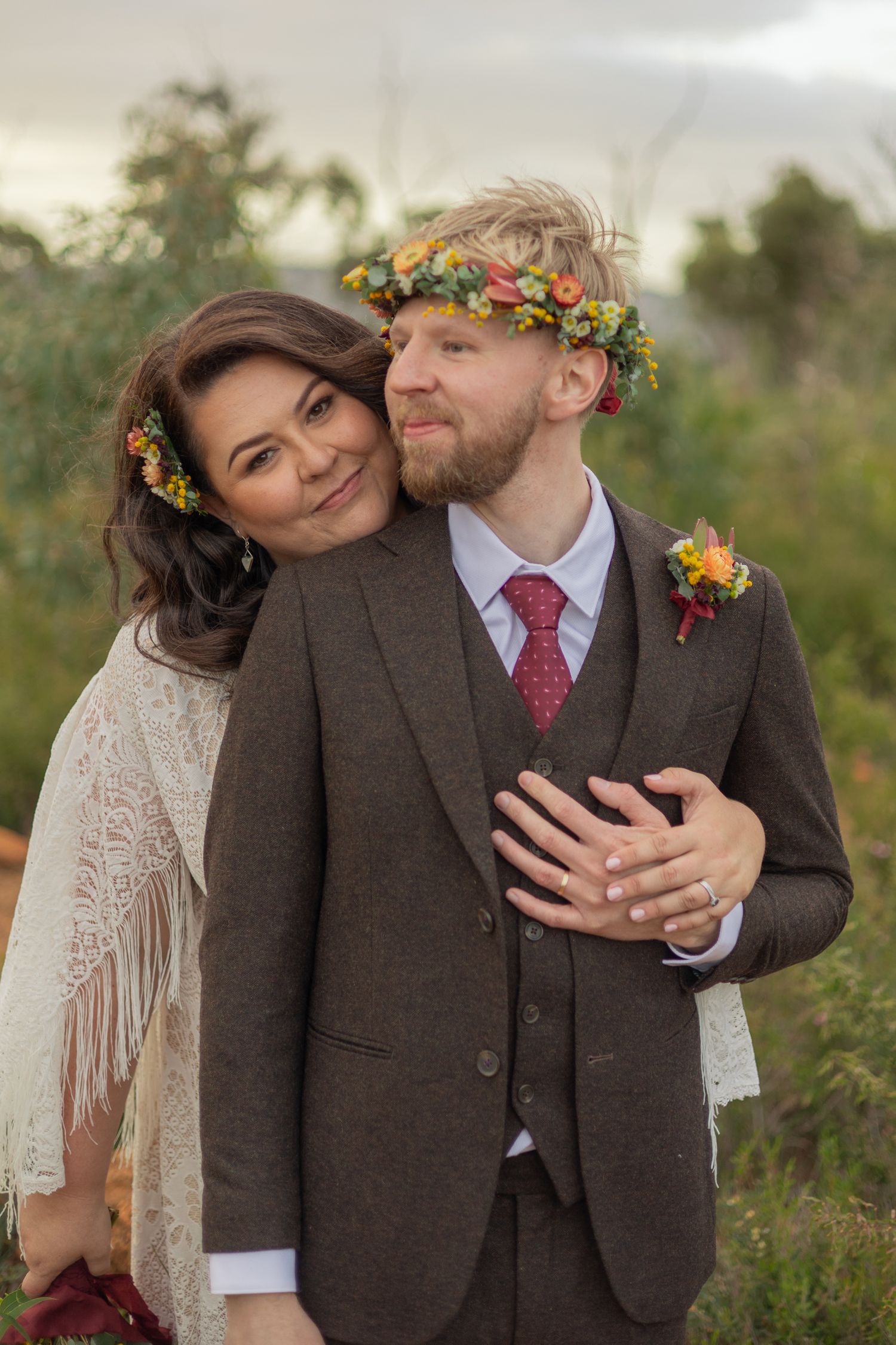 A couple in wedding attire share intimate moments in a field, with the groom wearing a brown suit and floral crown.