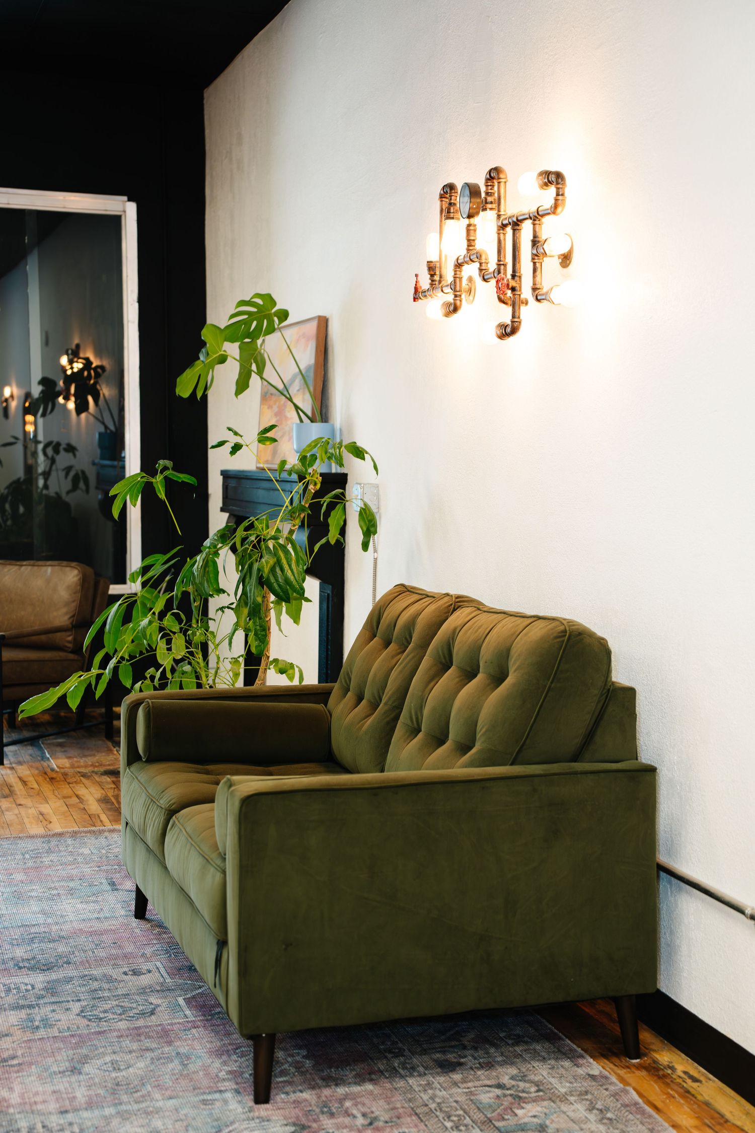 A green velvet tufted sofa with wooden legs sits against a white wall with potted plants and wall sconces ideal for event hosting.