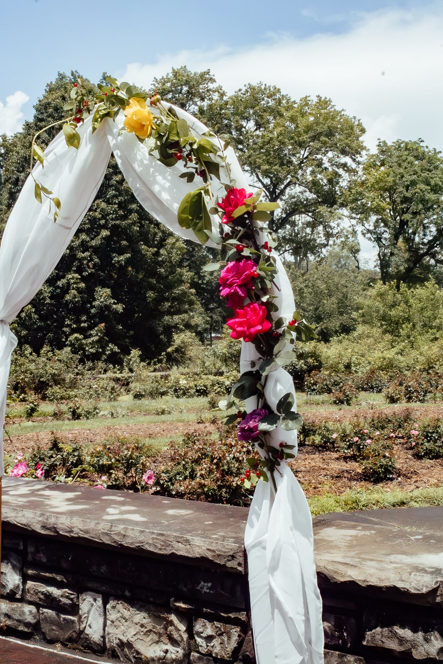 A wedding arch decorated with colorful flowers and white fabric against a garden backdrop.