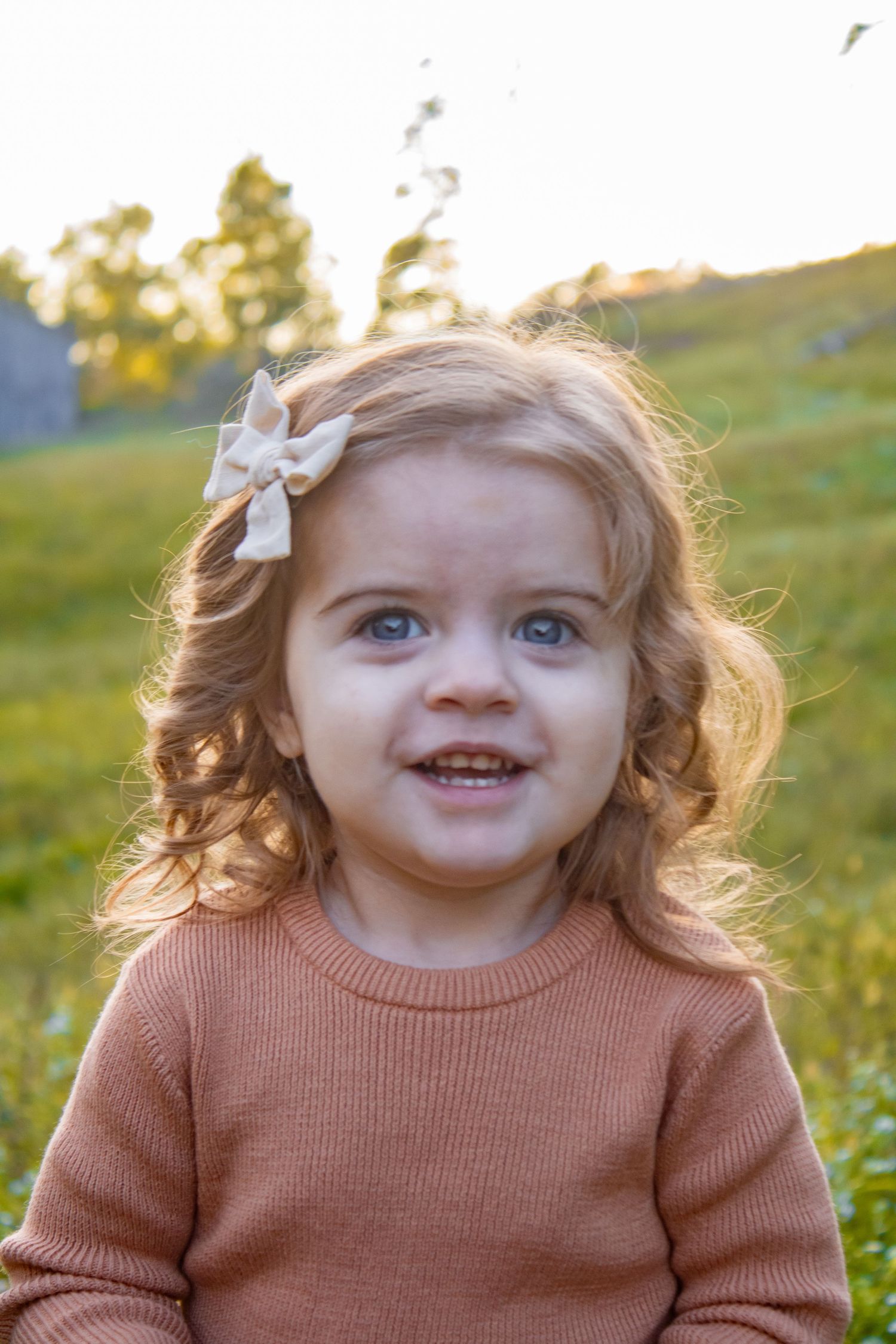 Young child in peach sweater and white hair bow smiles brightly in golden sunlight.