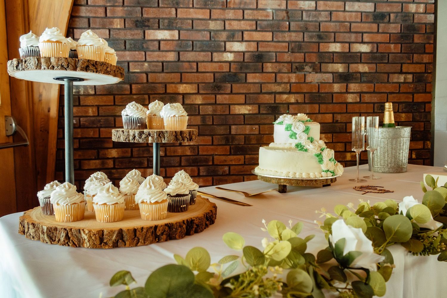 Rustic wedding dessert display featuring cupcakes and treats on wooden slices against a brick wall.