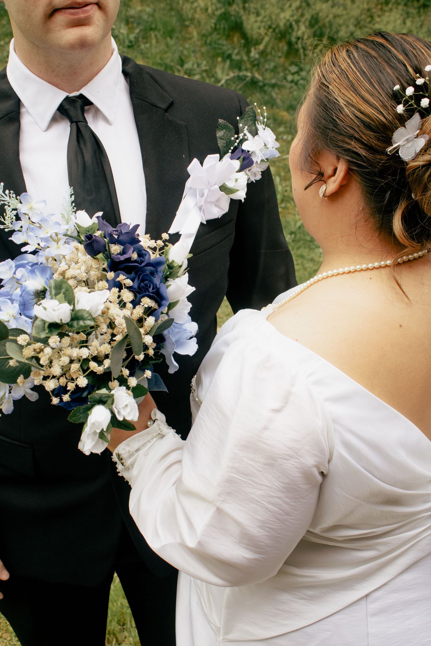 Wedding couple in formal attire pose with blue and white floral bouquet outdoors.