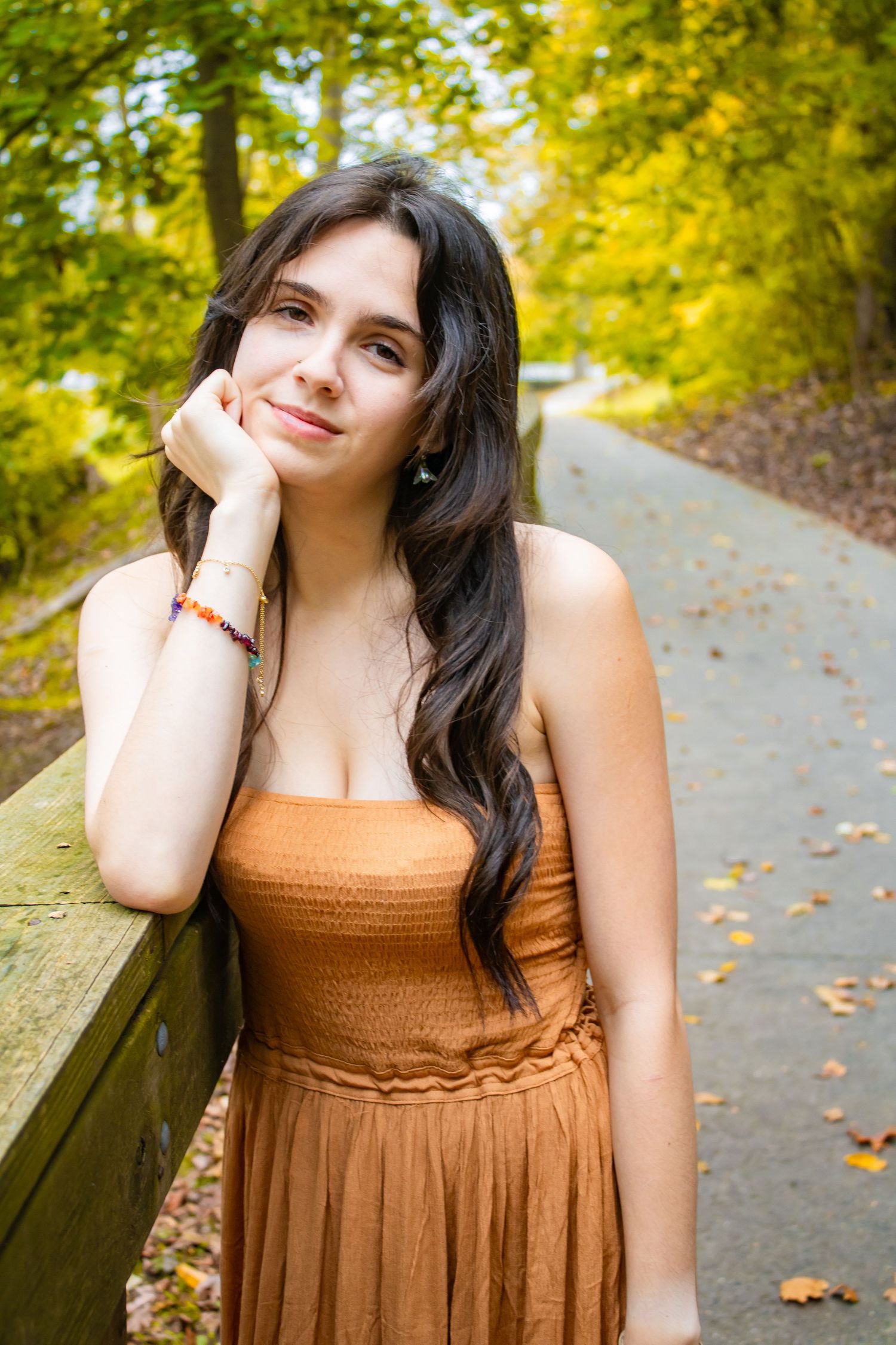 Young woman in brown dress poses on wooden bench surrounded by autumn foliage.