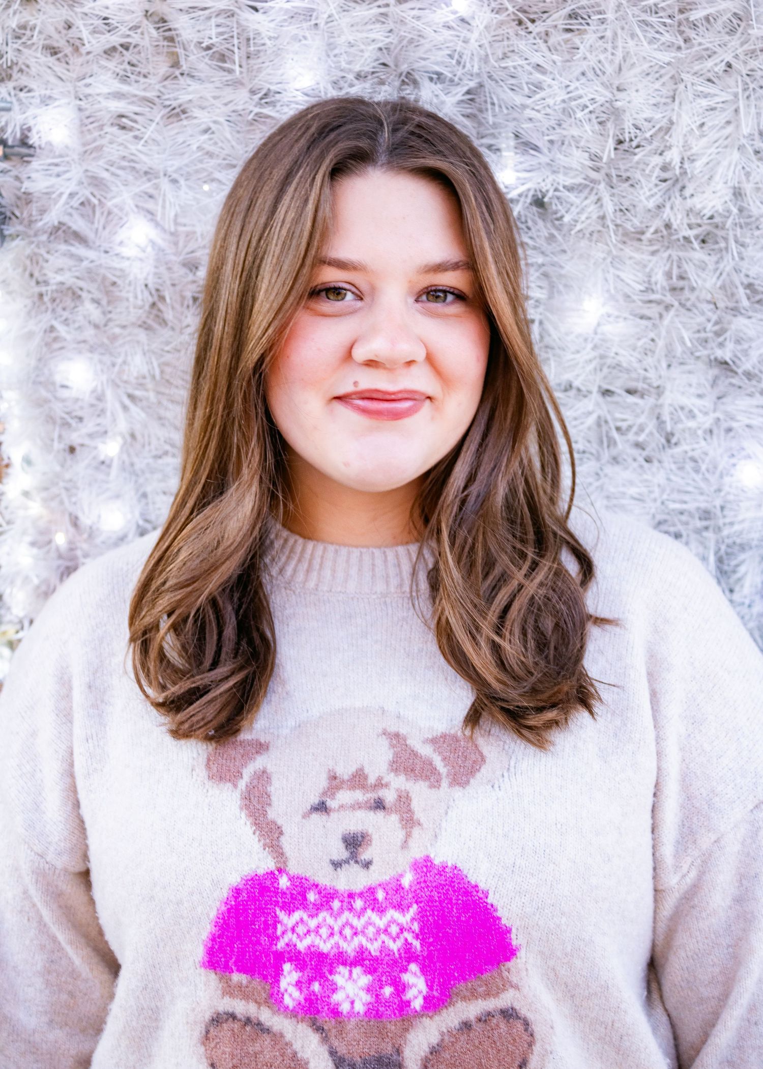 A person wearing a cream colored sweater with a teddy bear design smiles warmly against a white textured background.