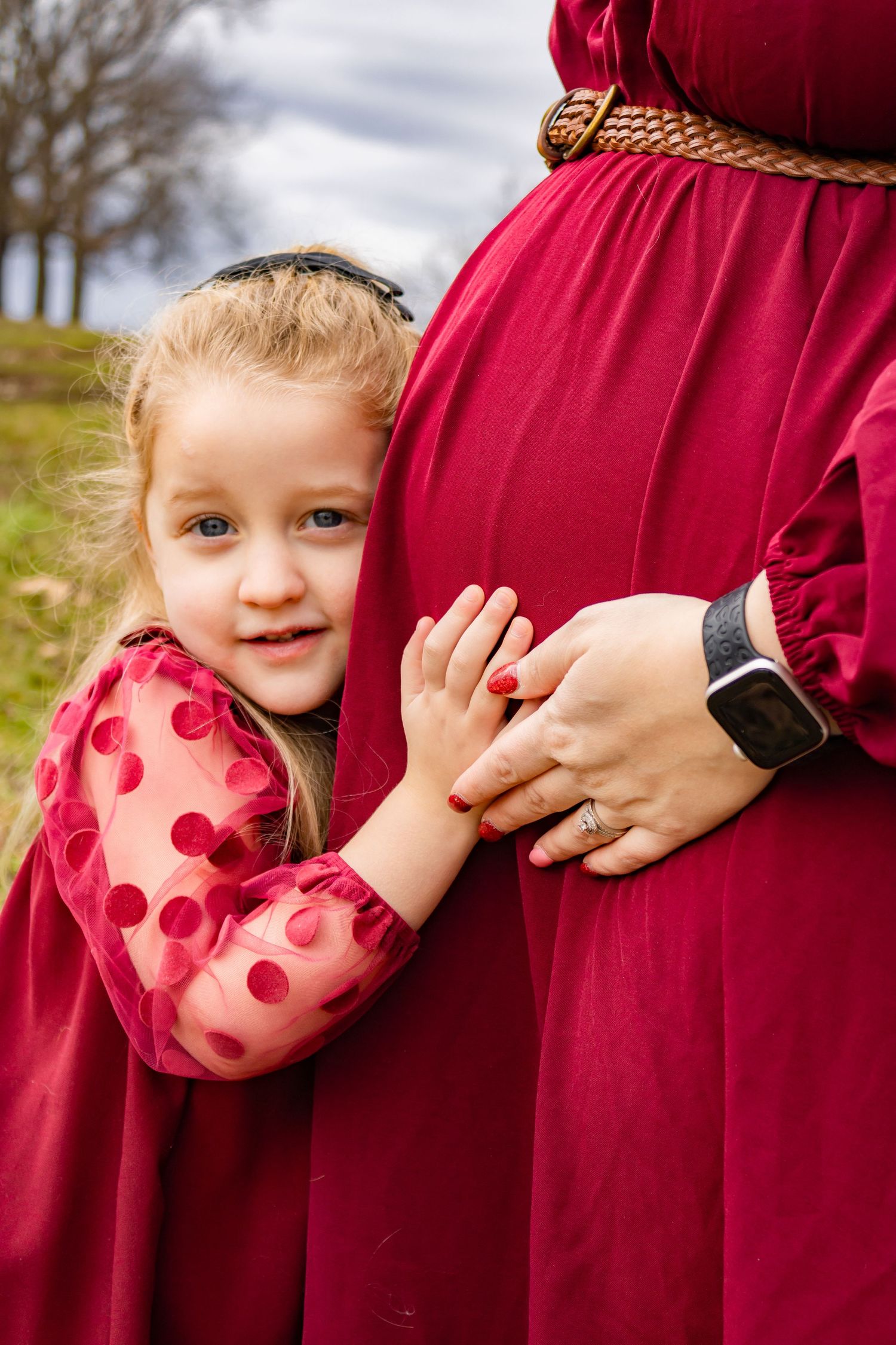 Child in a pink polka dot shirt peeks out from behind a red maternity dress in a sweet outdoor photo series.