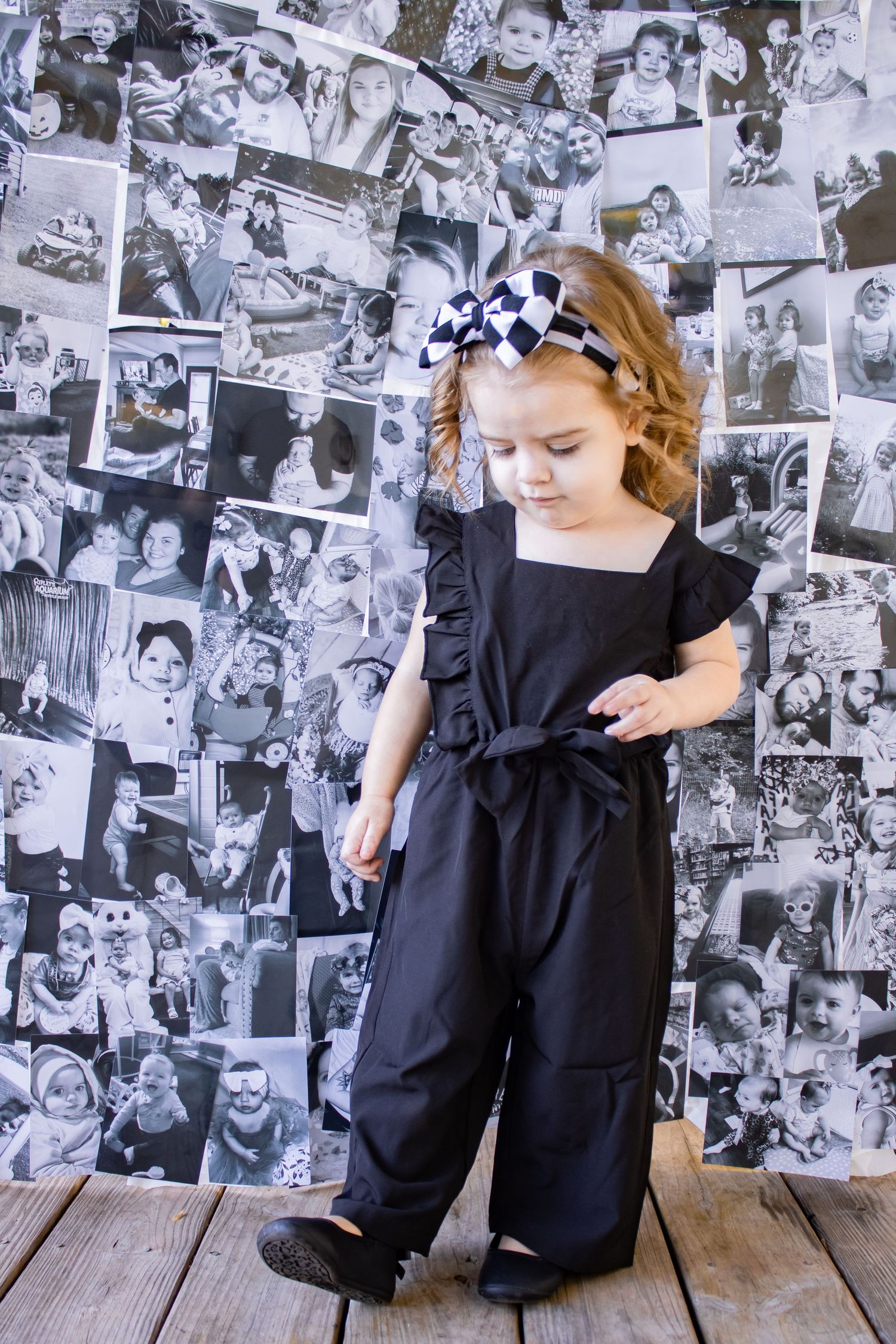 Child in black dress sits on wooden floor against wall covered in vintage black and white photographs.