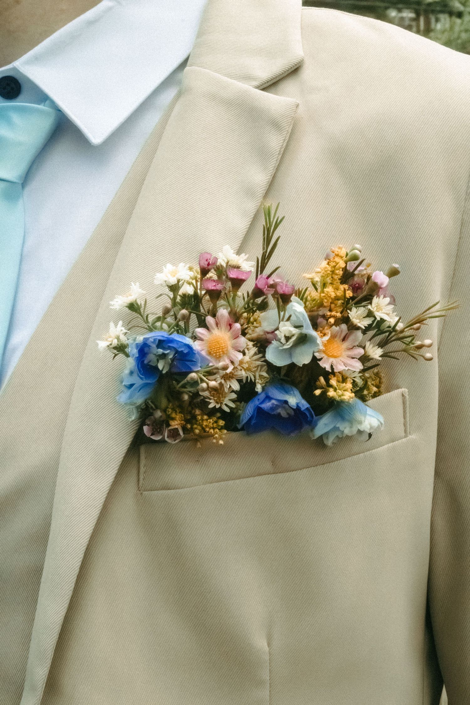 Delicate floral boutonniere with blue and pink flowers pinned to a beige suit lapel.