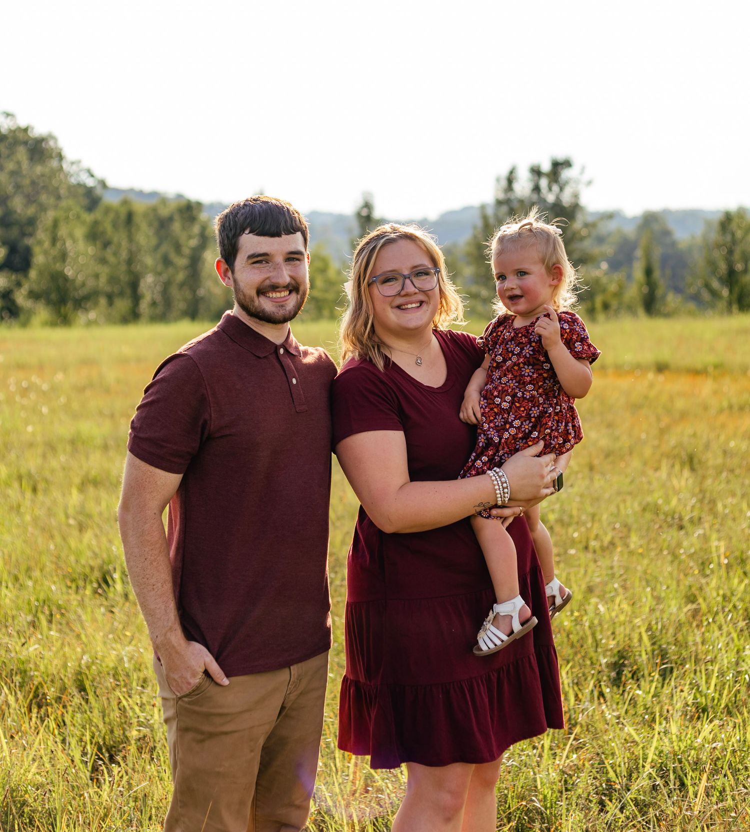 Family wearing burgundy outfits poses together in a sunny golden field during sunset.
