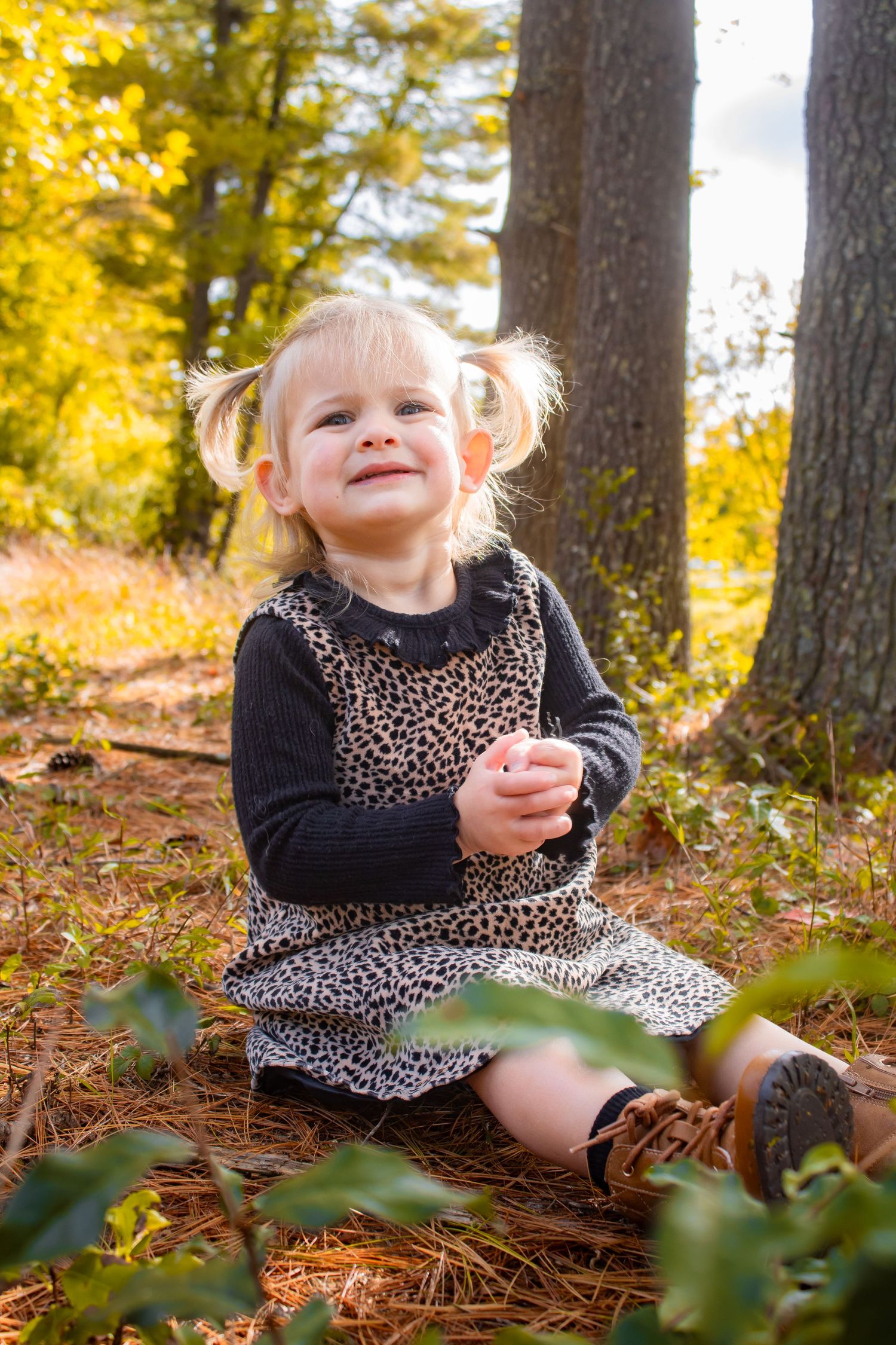 Child in leopard print dress sitting among fallen pine needles in autumn forest.