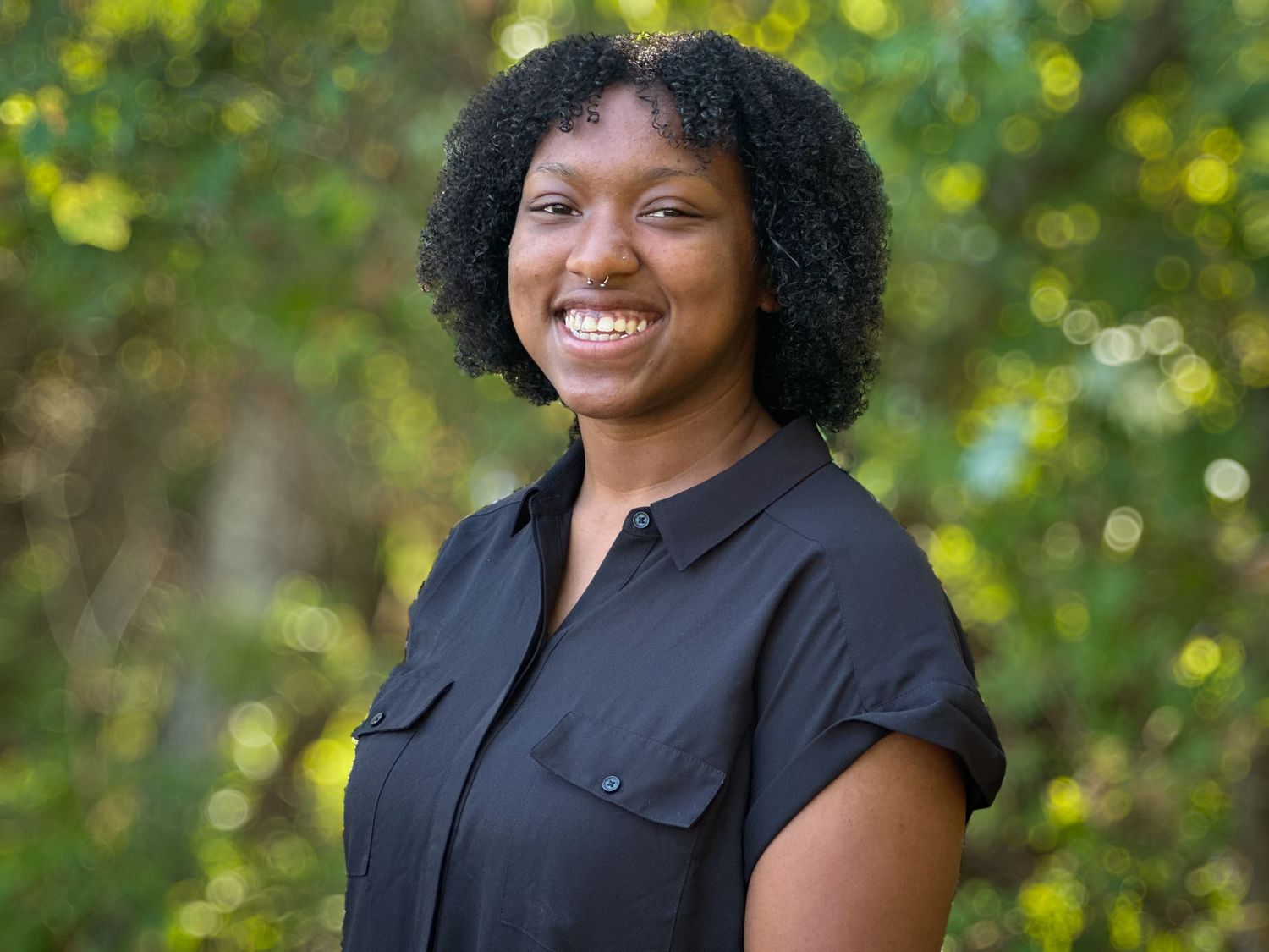 Professional headshot of person in black top against green natural background.