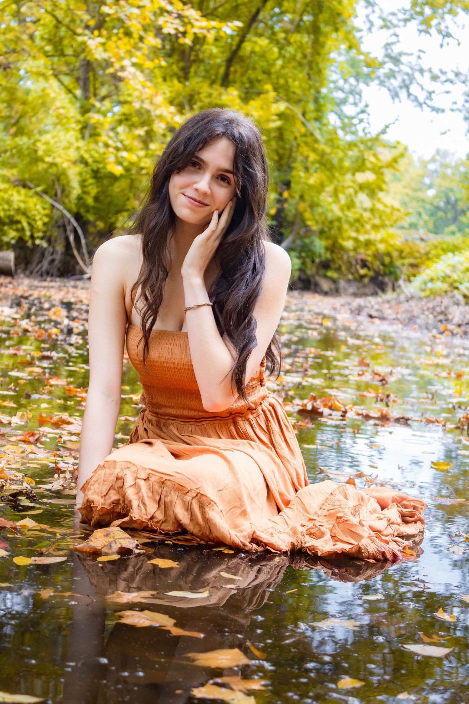 Model in rust-colored dress poses artistically by water with fallen leaves and autumn foliage.