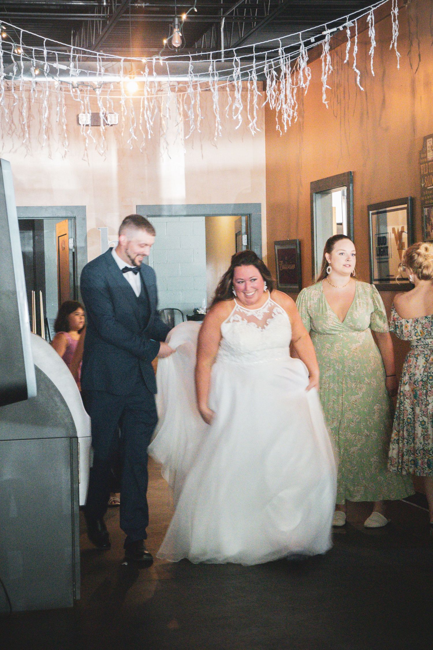 Wedding party dancing and celebrating under hanging lights at an indoor reception.