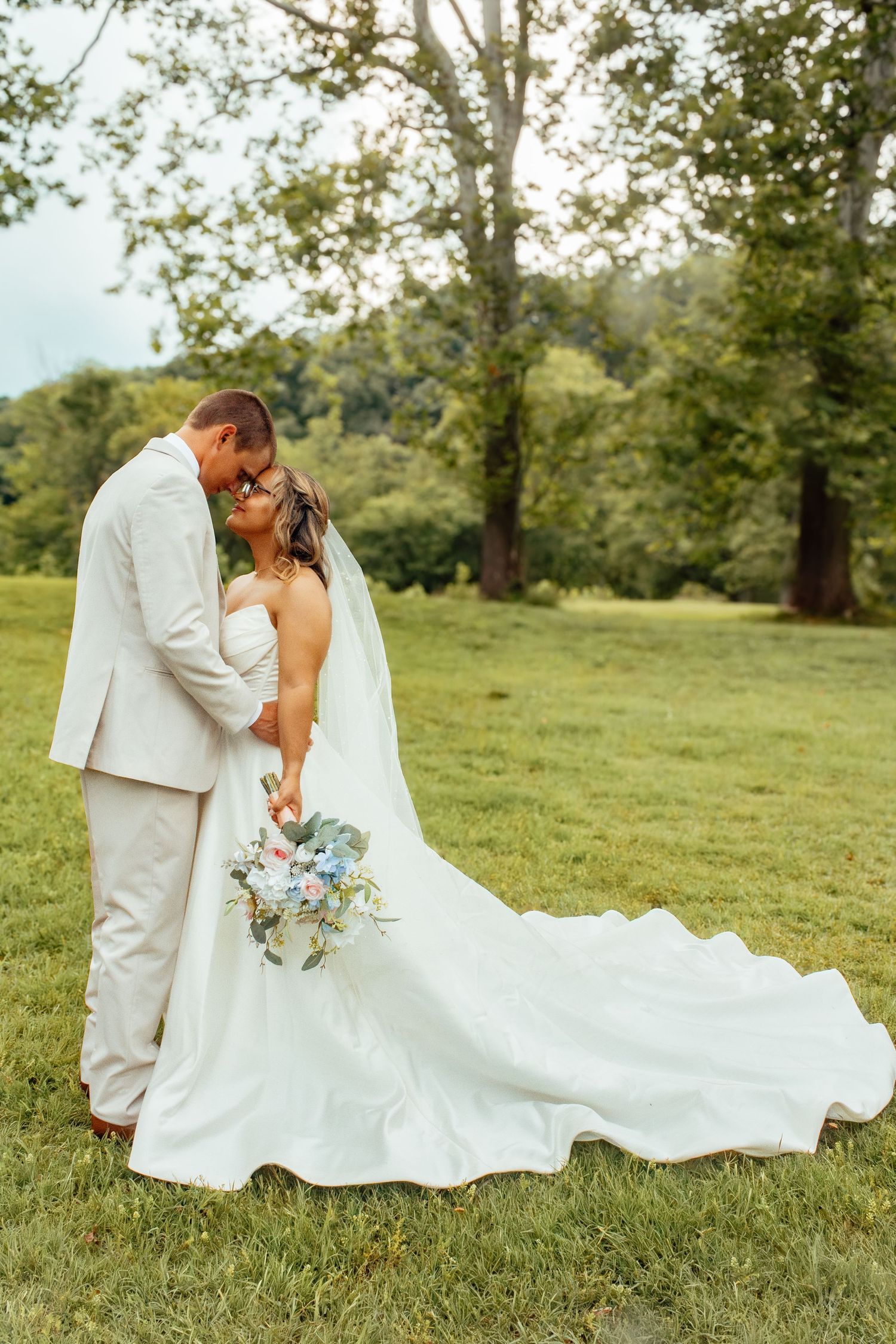 A wedding couple shares intimate moments on a grassy hillside surrounded by trees.