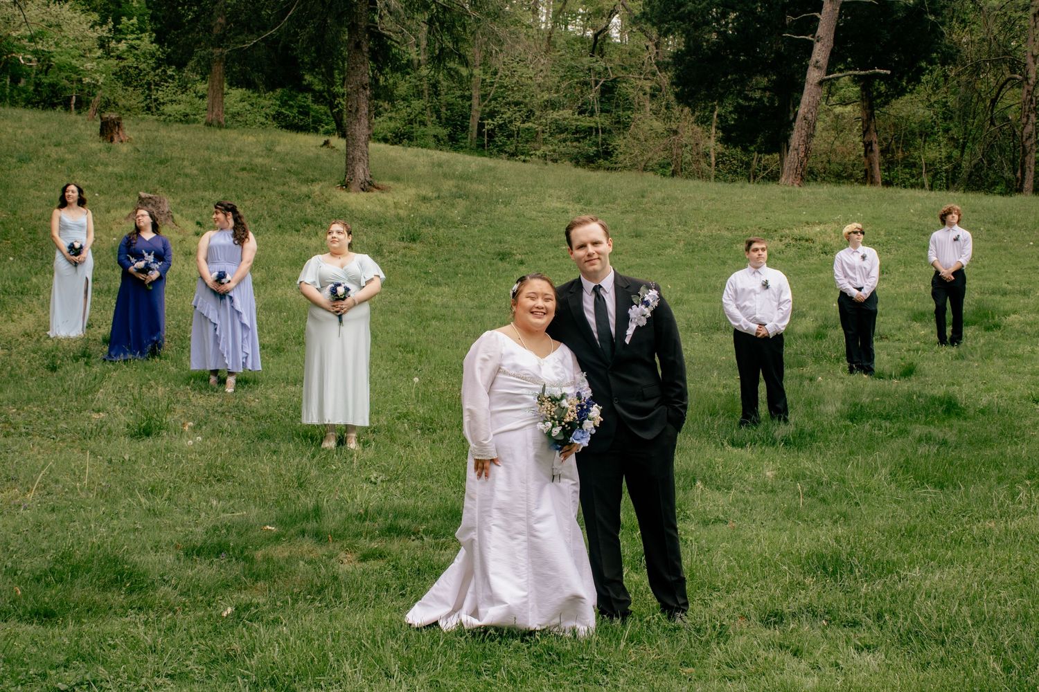Wedding party poses for photos on a grassy hillside with trees in the background.