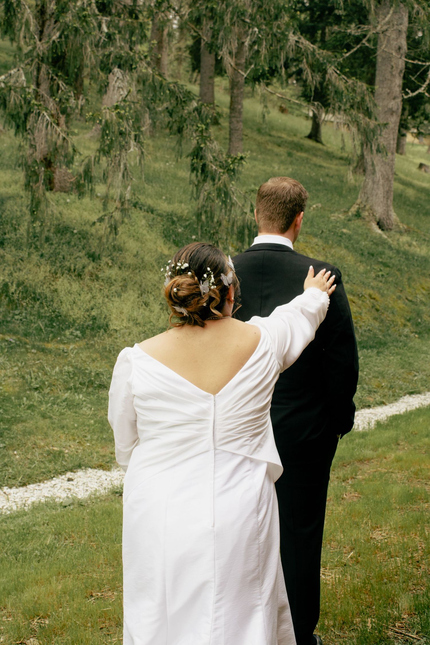 A couple in wedding attire walk along a scenic outdoor path surrounded by trees.