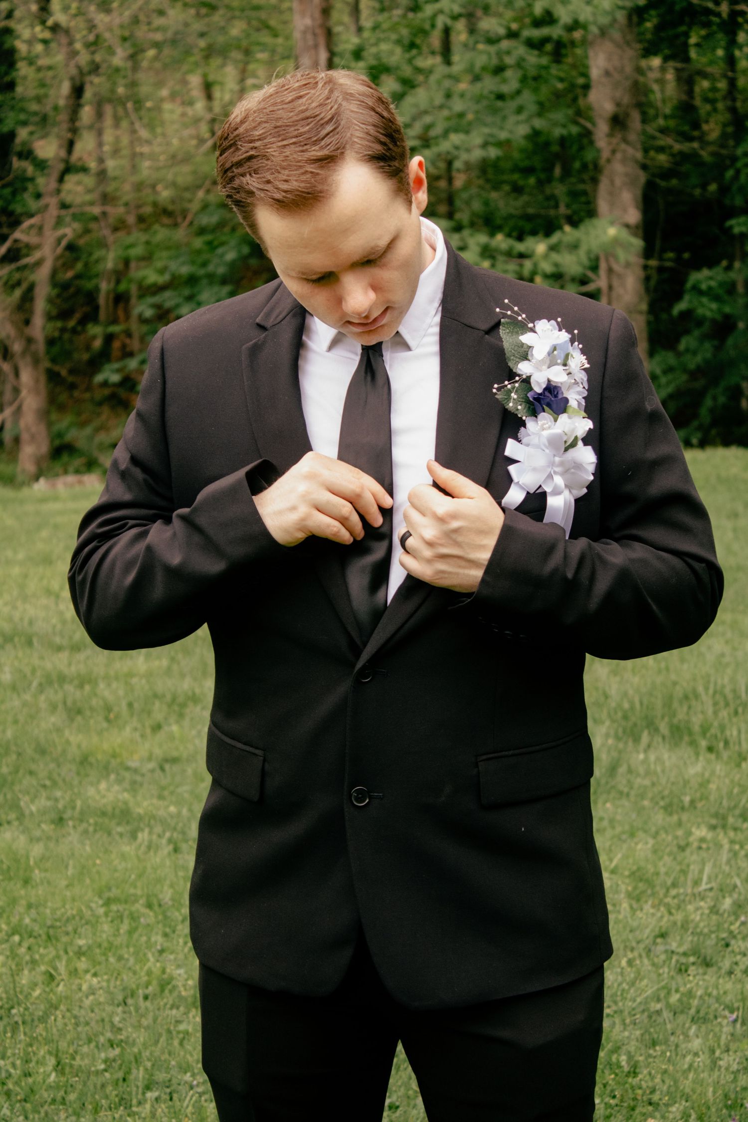 Person in black suit adjusts their tie while standing in a green outdoor setting.