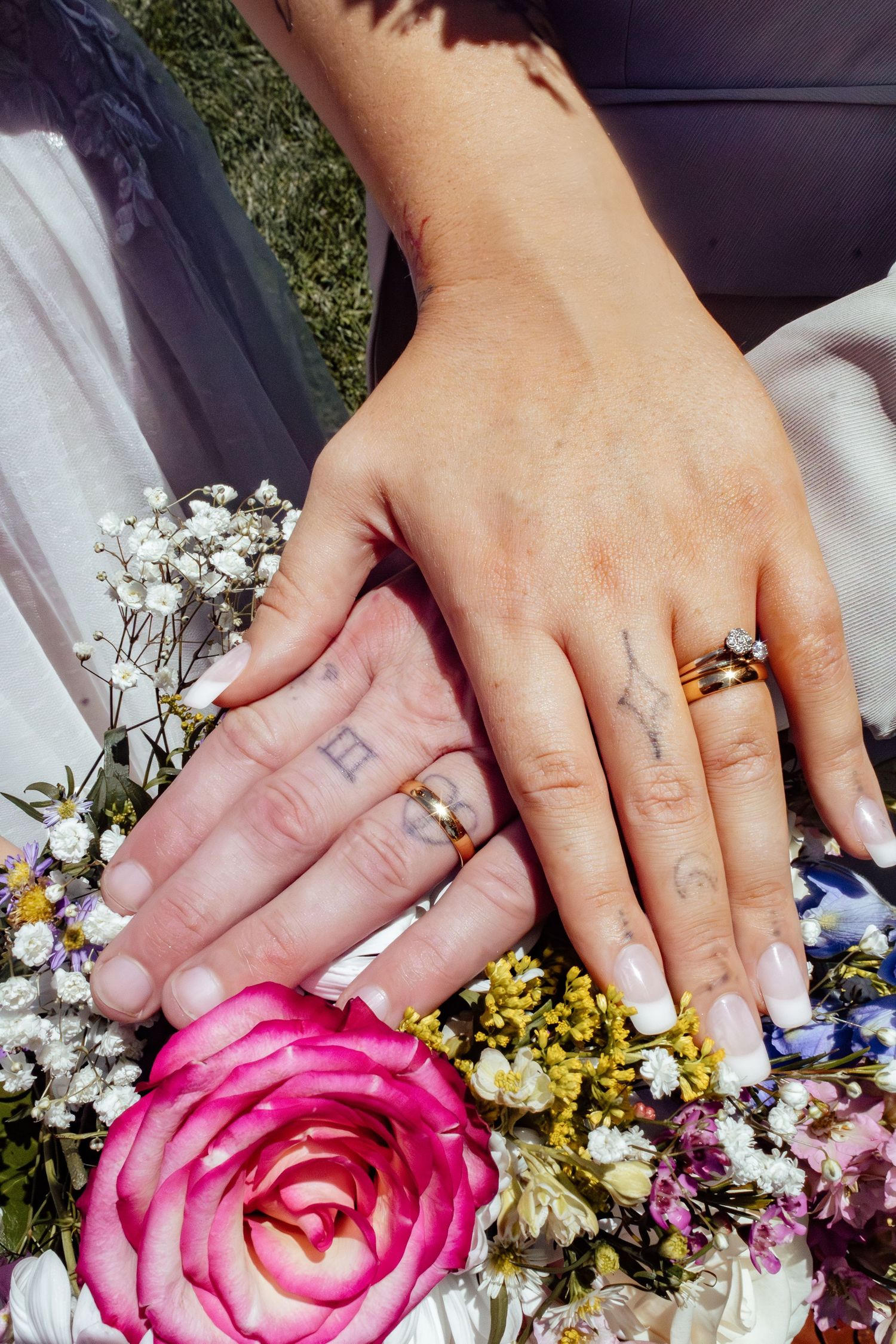 Wedding rings displayed with hands on a colorful floral bouquet featuring a pink rose.