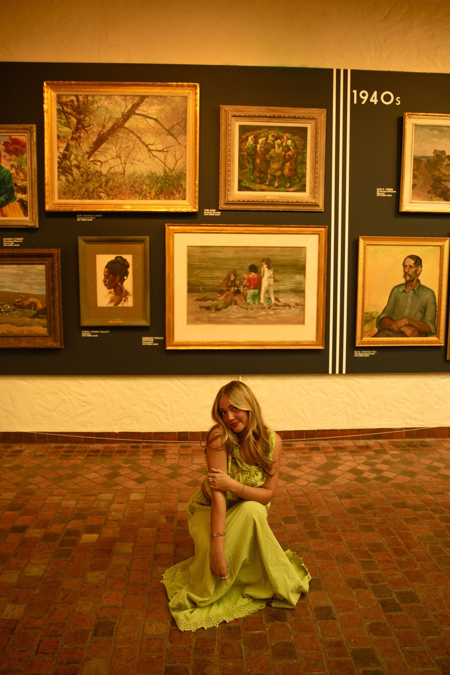 Woman in green dress sits on brick floor before 1940s art gallery wall displaying vintage framed paintings.
