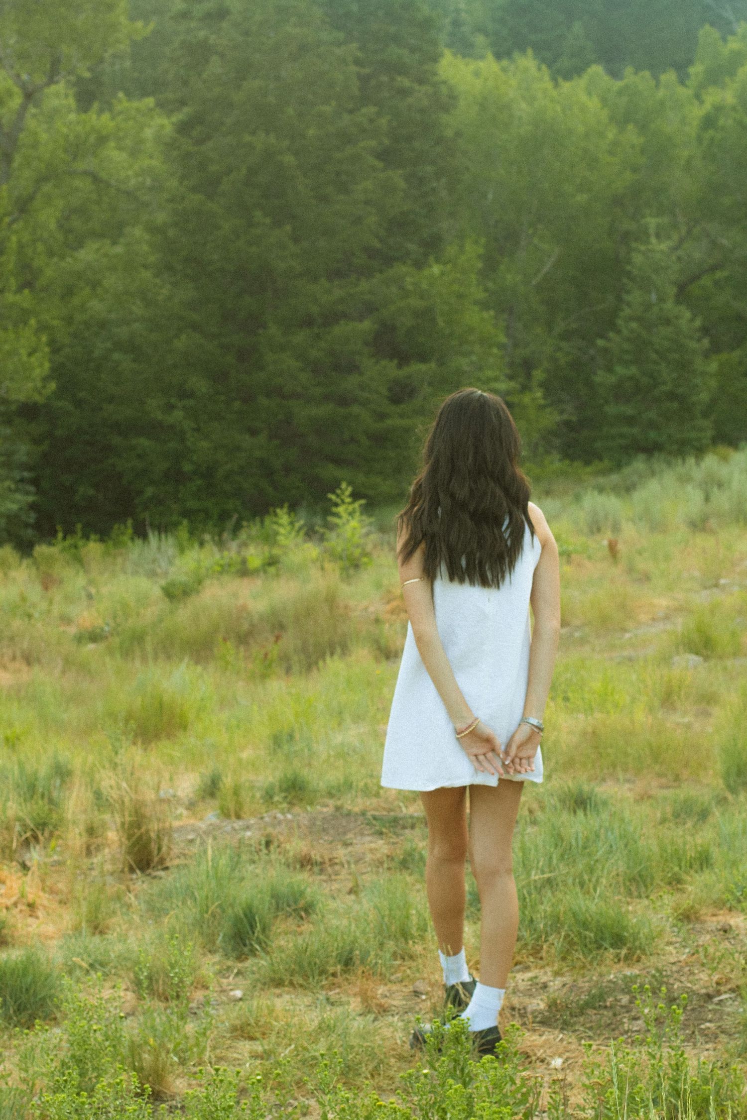 A person in a white dress walks through a grassy meadow with trees in the background.