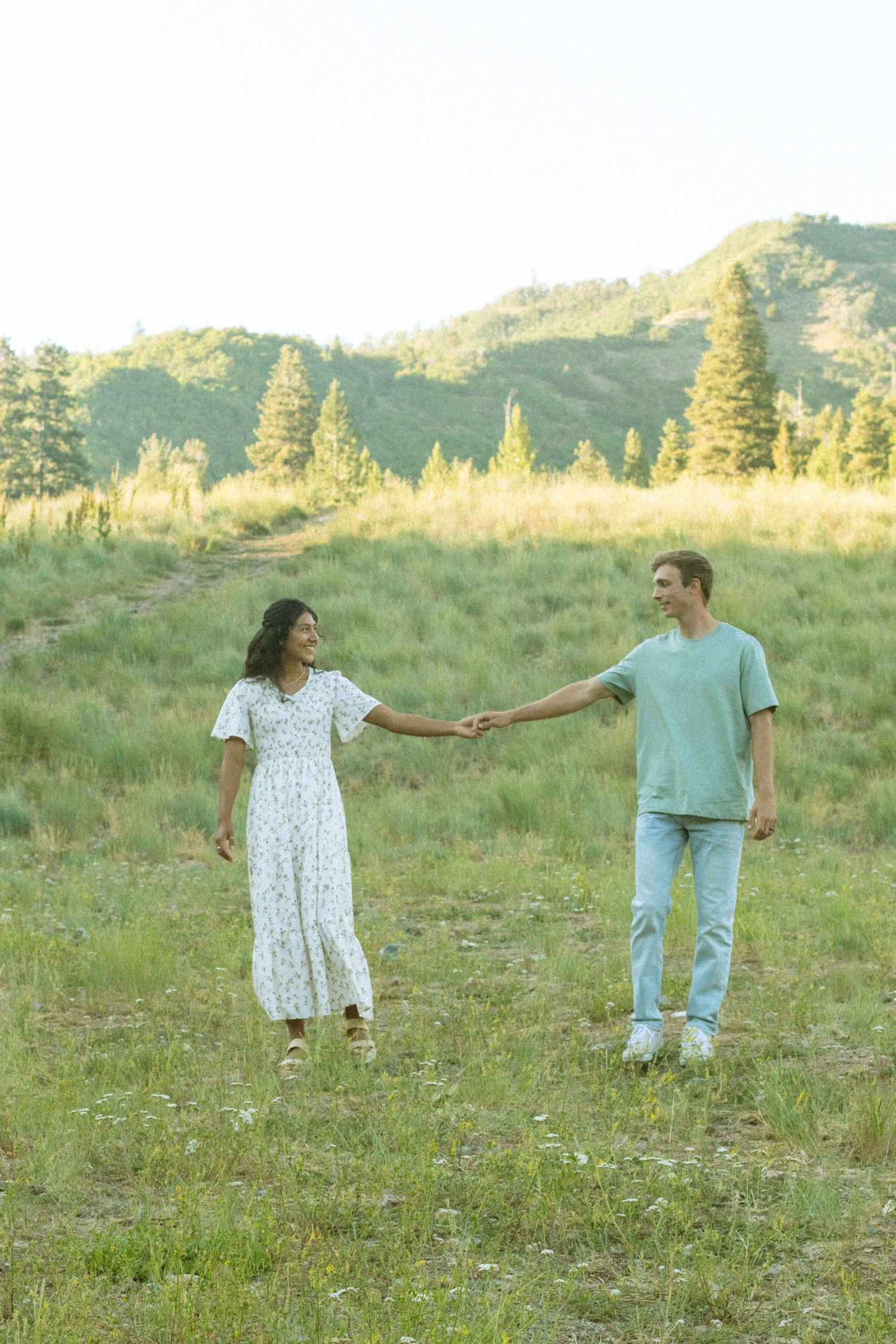 A couple holds hands while walking through a sunny mountain meadow in casual summer attire.
