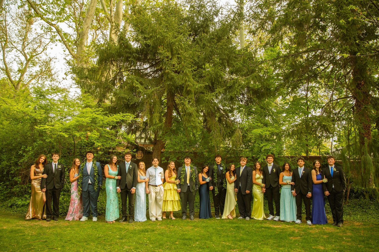 Large group of teens in formal prom attire posing together on a green lawn surrounded by lush trees.