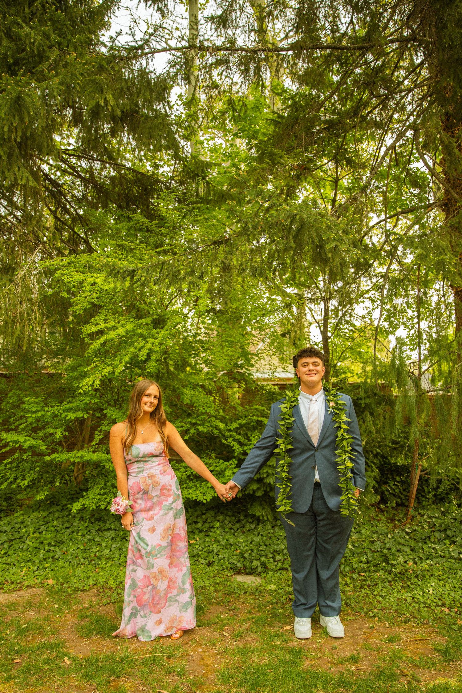 Young couple in formal wear holding hands outdoors, girl in floral dress, boy in blue suit, lush green trees background.