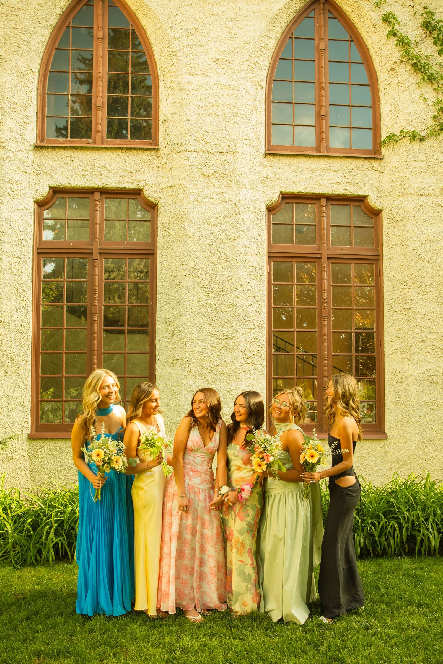 Six bridesmaids in colorful mismatched gowns holding floral bouquets stand laughing outside a stone chapel.