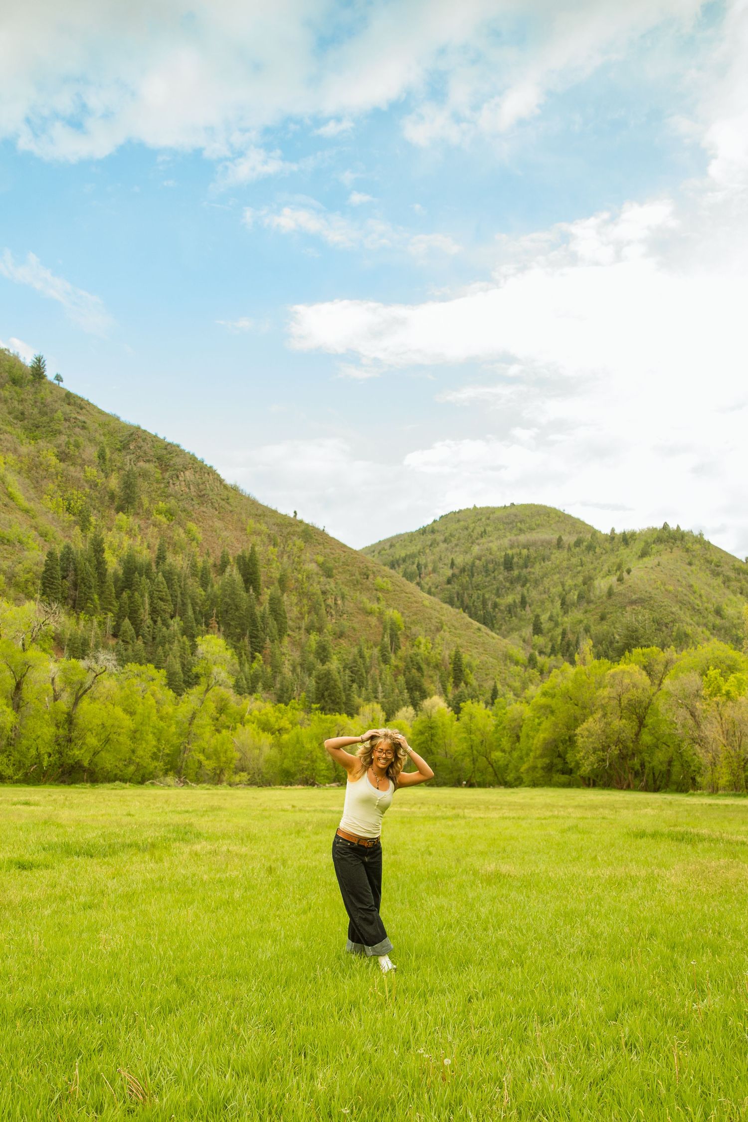 A young woman stands in a lush green meadow with forested mountains and blue sky in the background.