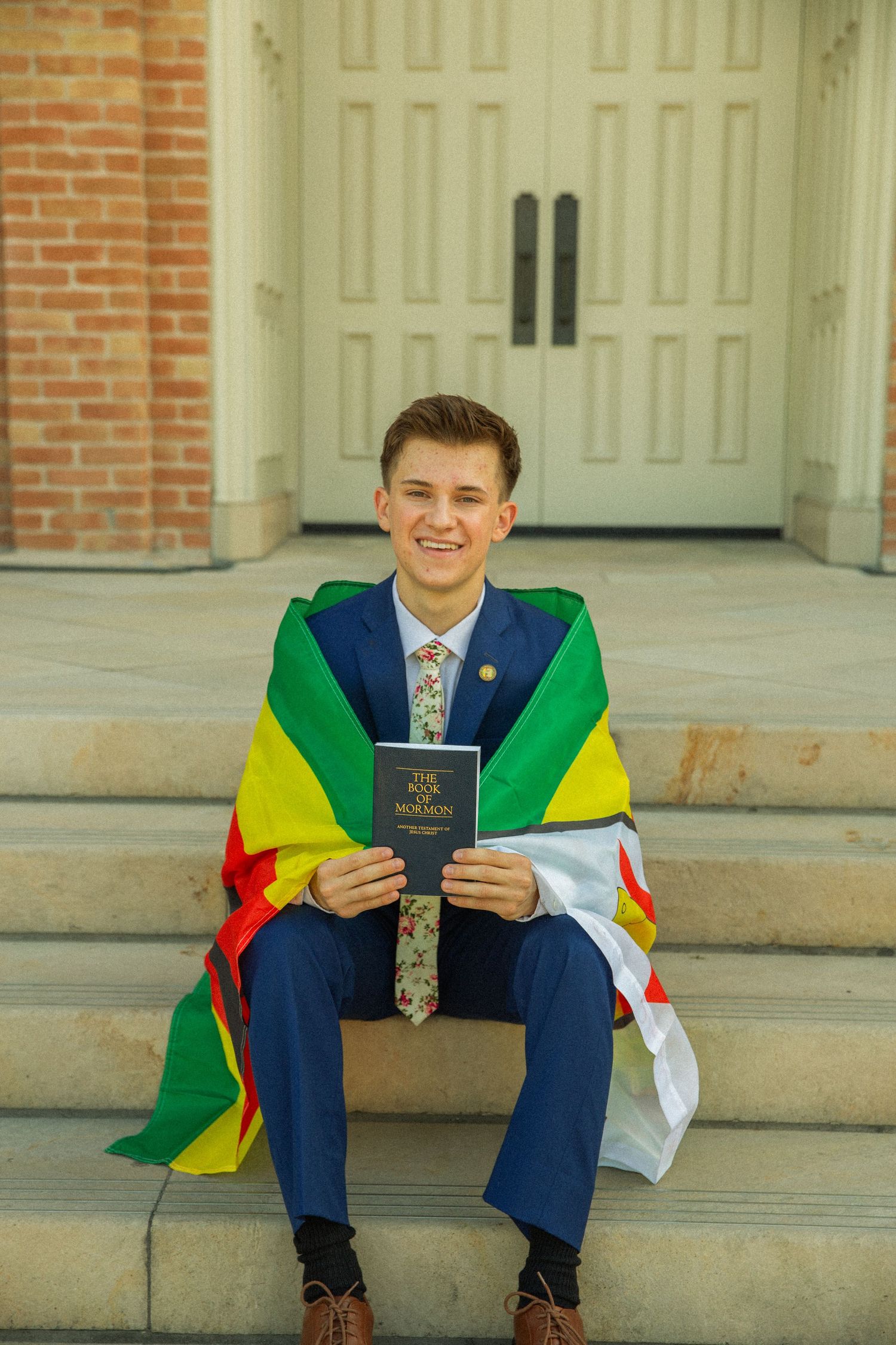 A missionary in a navy suit sits on stone steps holding a book while draped in a colorful South American flag.
