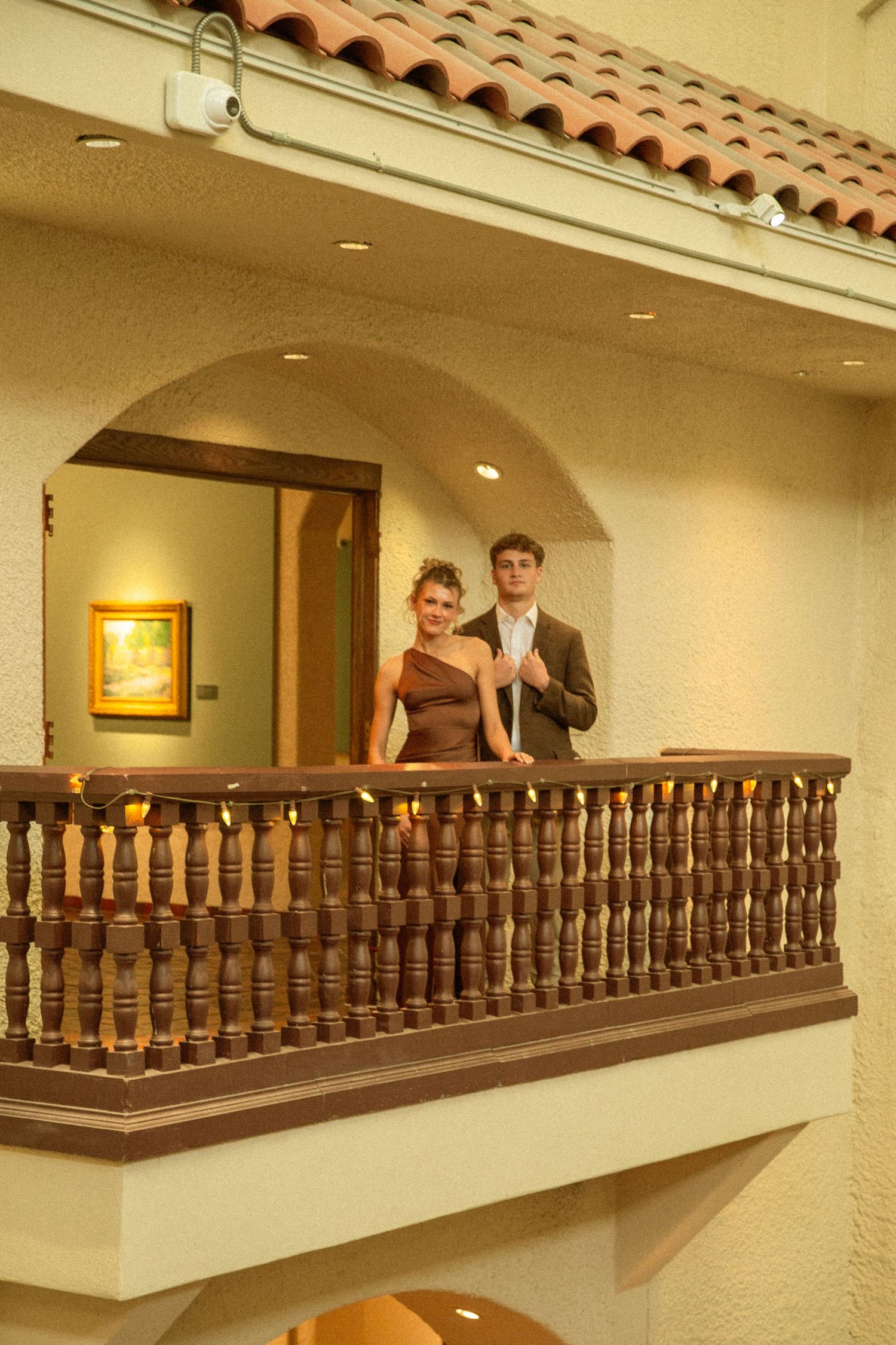 A couple stands at an elegant wooden balcony railing inside a Spanish-style building with warm lighting and terracotta tiles.