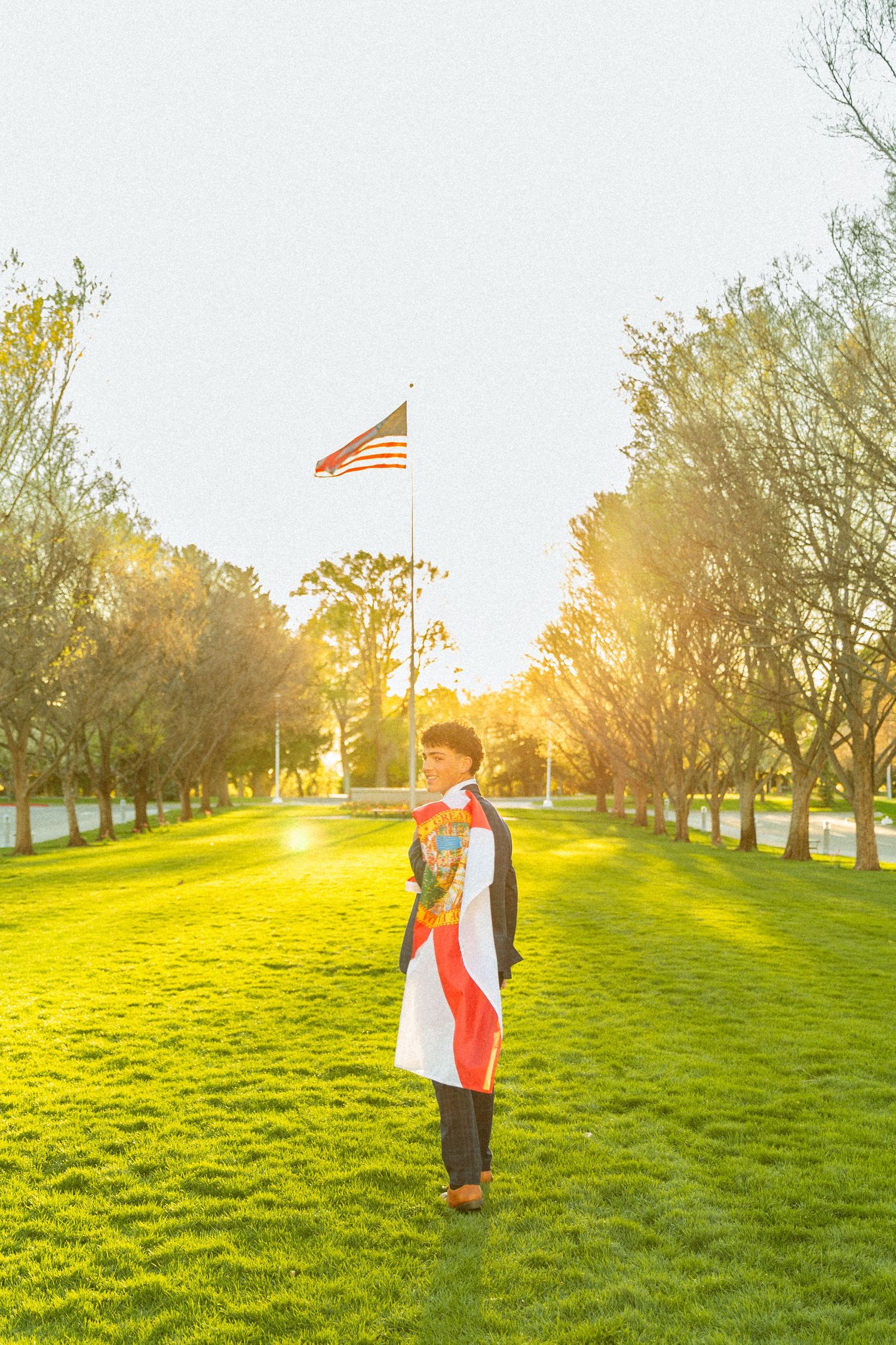 Person wrapped in colorful flag standing on green grass in park with American flag and sunlit trees in background.