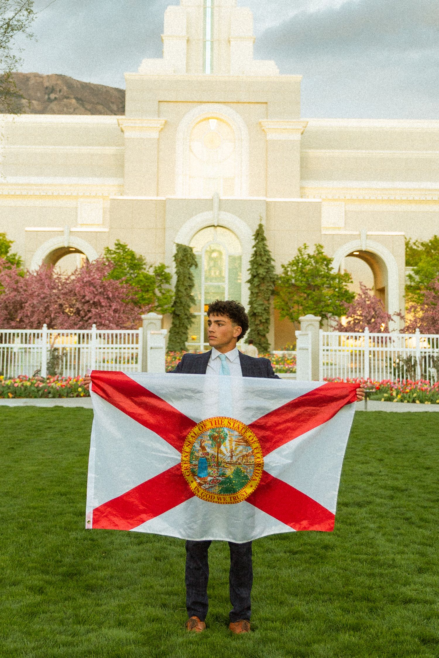 Young man in suit holding Florida state flag outdoors in front of a white temple with blooming trees.