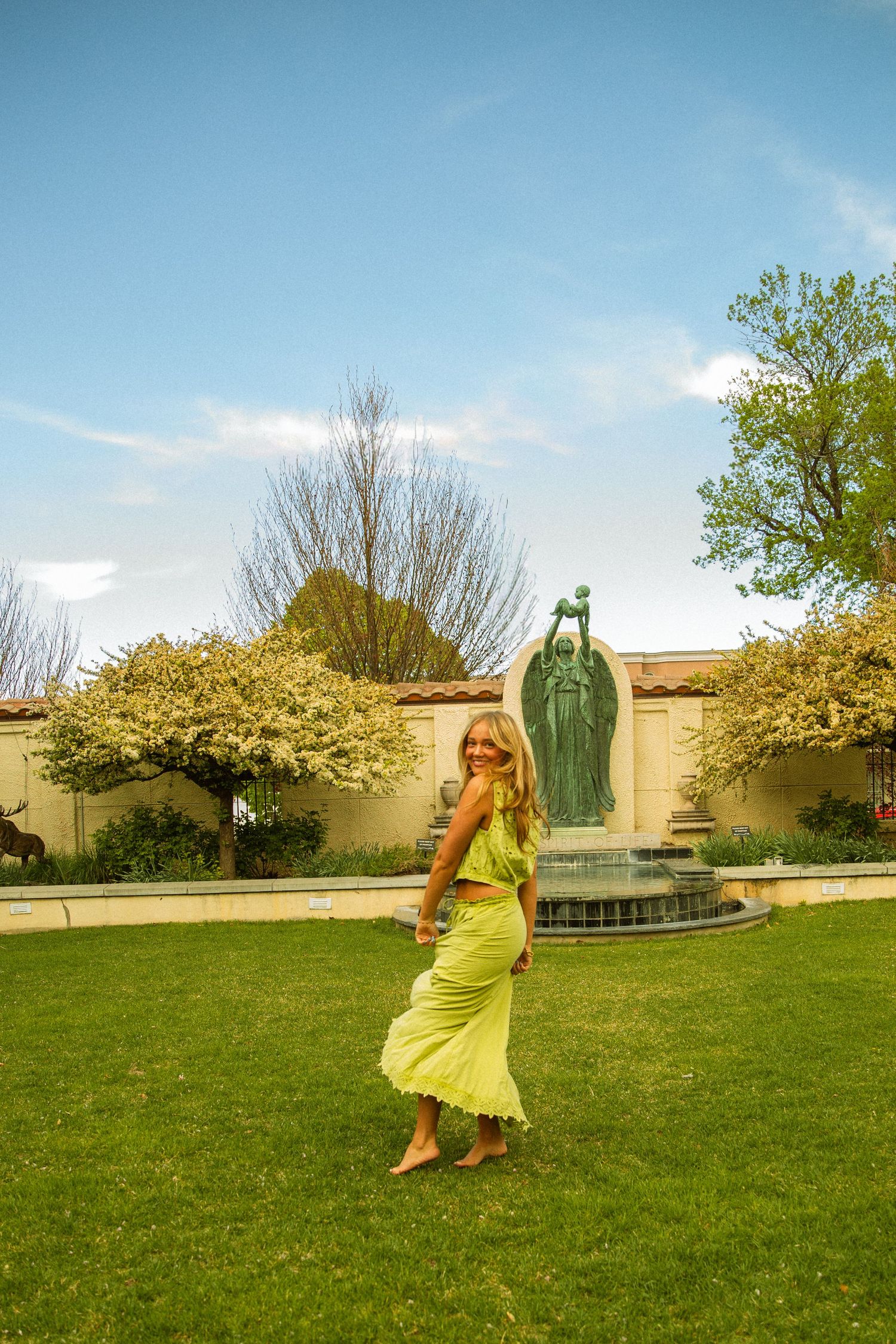 Woman in yellow-green outfit dancing on green lawn near ornate fountain statue with blooming trees and blue sky.