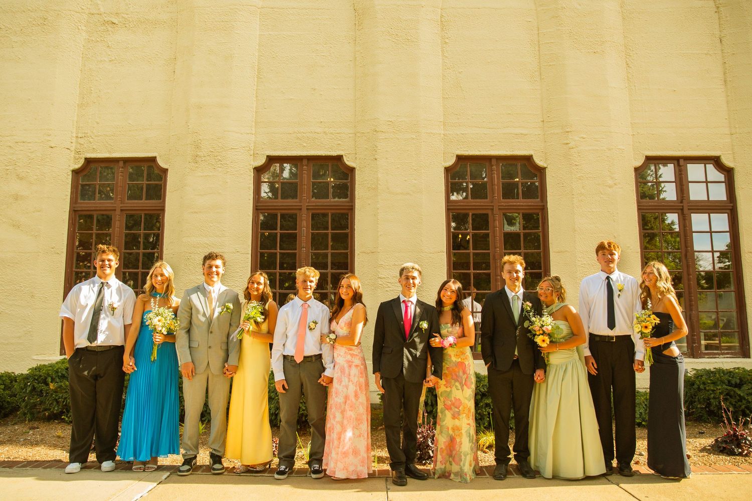 Group of prom couples posing in formal attire outside a classic building with large windows.